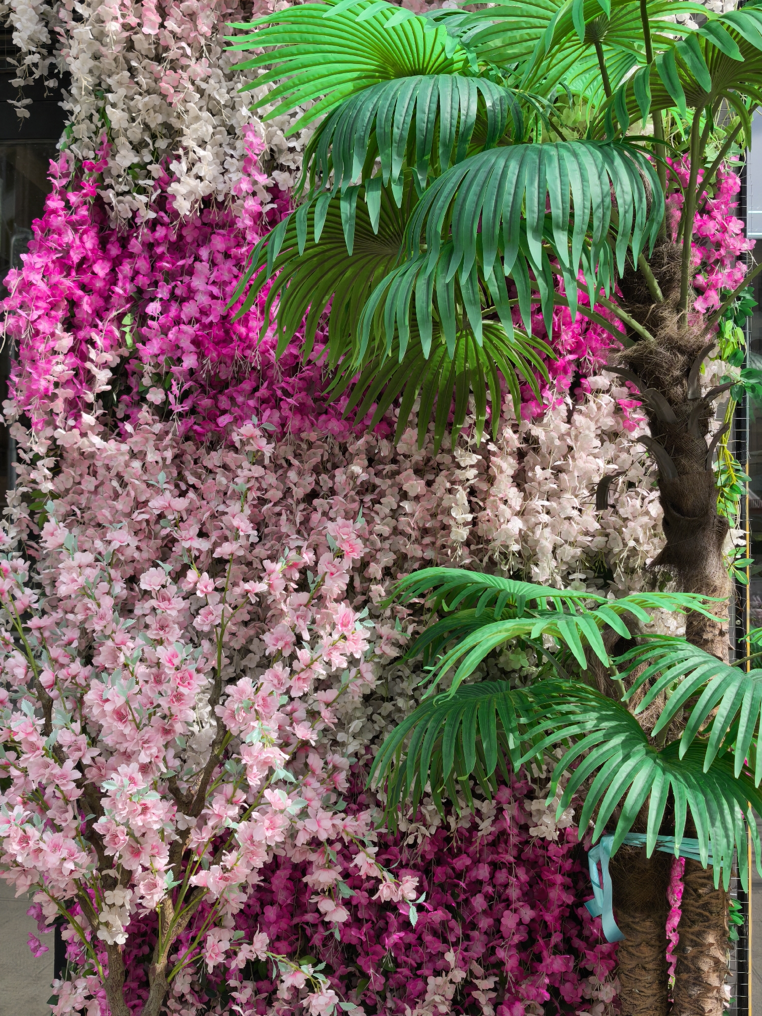 This image showcases a vibrant, dense decorative display composed entirely of artificial flora, likely situated indoors or in a sheltered urban environment such as Moscow, Russia. The scene is dominated by a rich tapestry of synthetic flowers and palm trees, creating a lush, tropical-floral wall.
On the left side, the background is covered by abundant cascades of small, faux flowers. These appear in layers, with a bright fuchsia-pink at the bottom and top, transitioning to lighter pink and white clusters in the middle. Interspersed among these hanging blossoms, especially in the lower left quadrant, are branches of larger, pink cherry blossom-like flowers with small, pale green leaves, adding variety to the floral textures.
To the right, two prominent artificial palm trees rise from the bottom, extending upwards. They feature textured, fibrous brown trunks, one of which has a light blue ribbon tied around it. Large, bright green, segmented palm fronds fan out, partially obscuring the floral background and adding a strong verdant contrast to the pink and white flowers.
The overall impression is one of an elaborate, colorful, and highly maintained decorative installation, serving as a backdrop or accent feature. No people or text are visible, and the artificial nature of the plants suggests a controlled setting rather than a natural outdoor landscape.