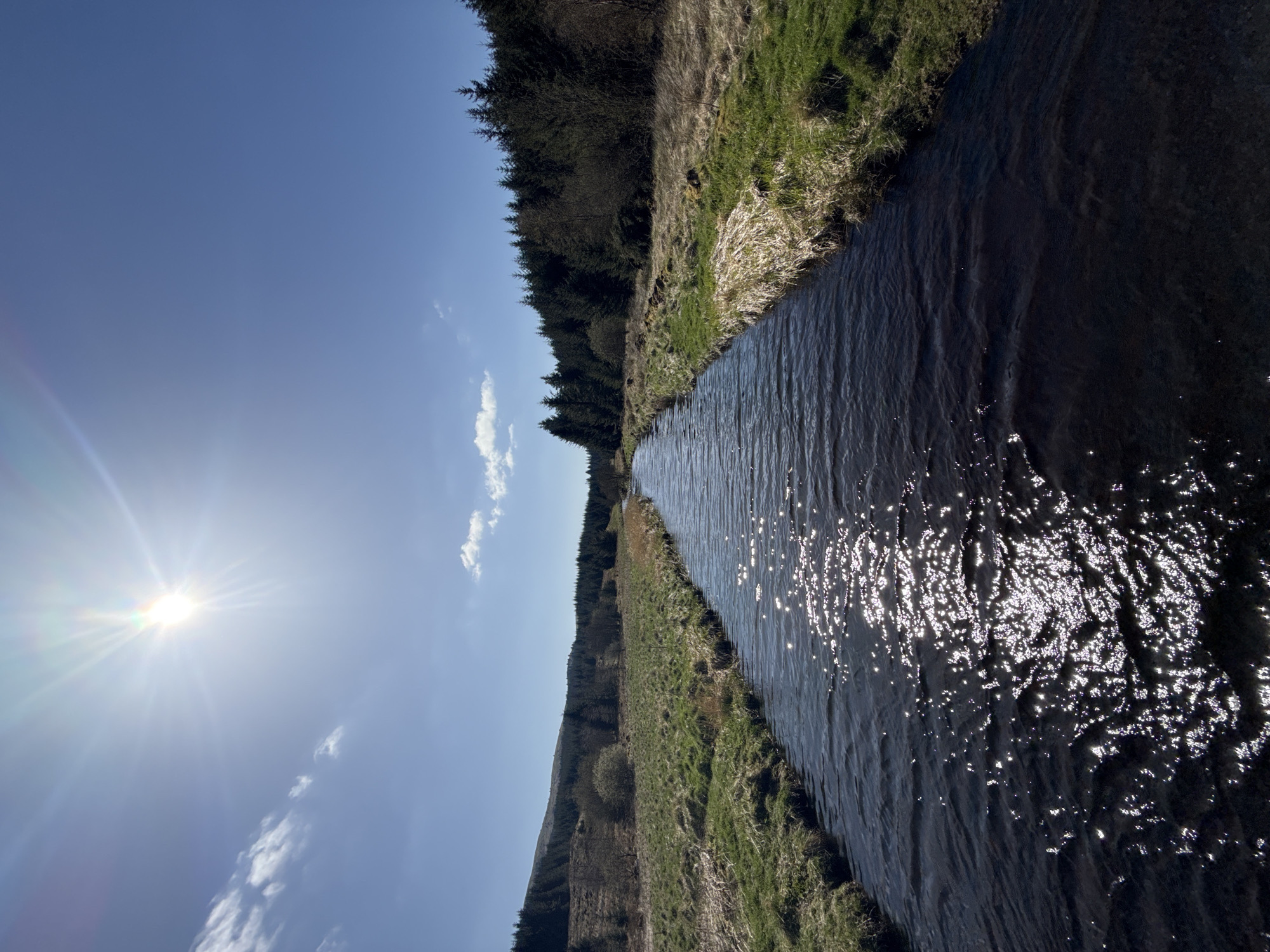 The image shows a narrow waterway running through a landscape. On one side, a line of trees runs along the bank. On the opposite side, a grassy bank slopes up. The waterway reflects the sunlight. The sky is blue with some clouds. The location is unknown.