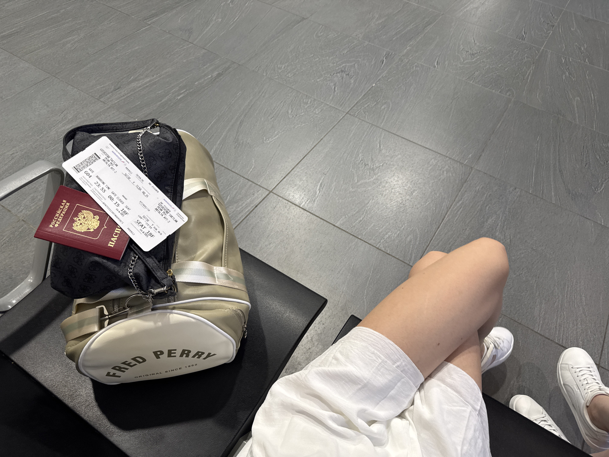 The image shows a passport and boarding pass on a bag in what appears to be an airport waiting area, likely at Bolshoye Savino Airport (USPP) in Bolshoye Savino, Russia. A person's legs, wearing white sneakers and a white skirt, are visible in the lower part of the frame, indicating they are seated. The passport is red with "РОССИЙСКАЯ ФЕДЕРАЦИЯ" (Russian Federation) printed in gold. The boarding pass is white and appears to be for a flight with information such as gate, boarding time, and seat number. The bag is a beige duffel bag with "FRED PERRY ORIGINAL SINCE 1952" printed on it, and a black patterned pouch is placed on top of it. The flooring is grey tile with a wood-like pattern.