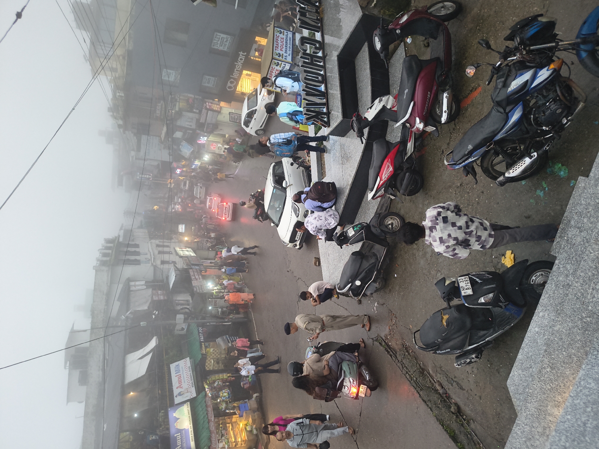 The image captures a street scene in Dharamshala, India, shrouded in dense fog, which obscures much of the background architecture. In the foreground, several parked scooters and motorcycles are visible, with a man in a checkered shirt walking away from the camera. A police officer is speaking to a couple on a scooter, while another person sits nearby. Further up the street, cars and more people are present, with shops lining both sides of the road. Signs for "Amul Milk" and "Lenskart" are discernible, indicating commercial activity. The wet road surface suggests recent rain. The overall atmosphere is hazy and somewhat muted due to the fog.