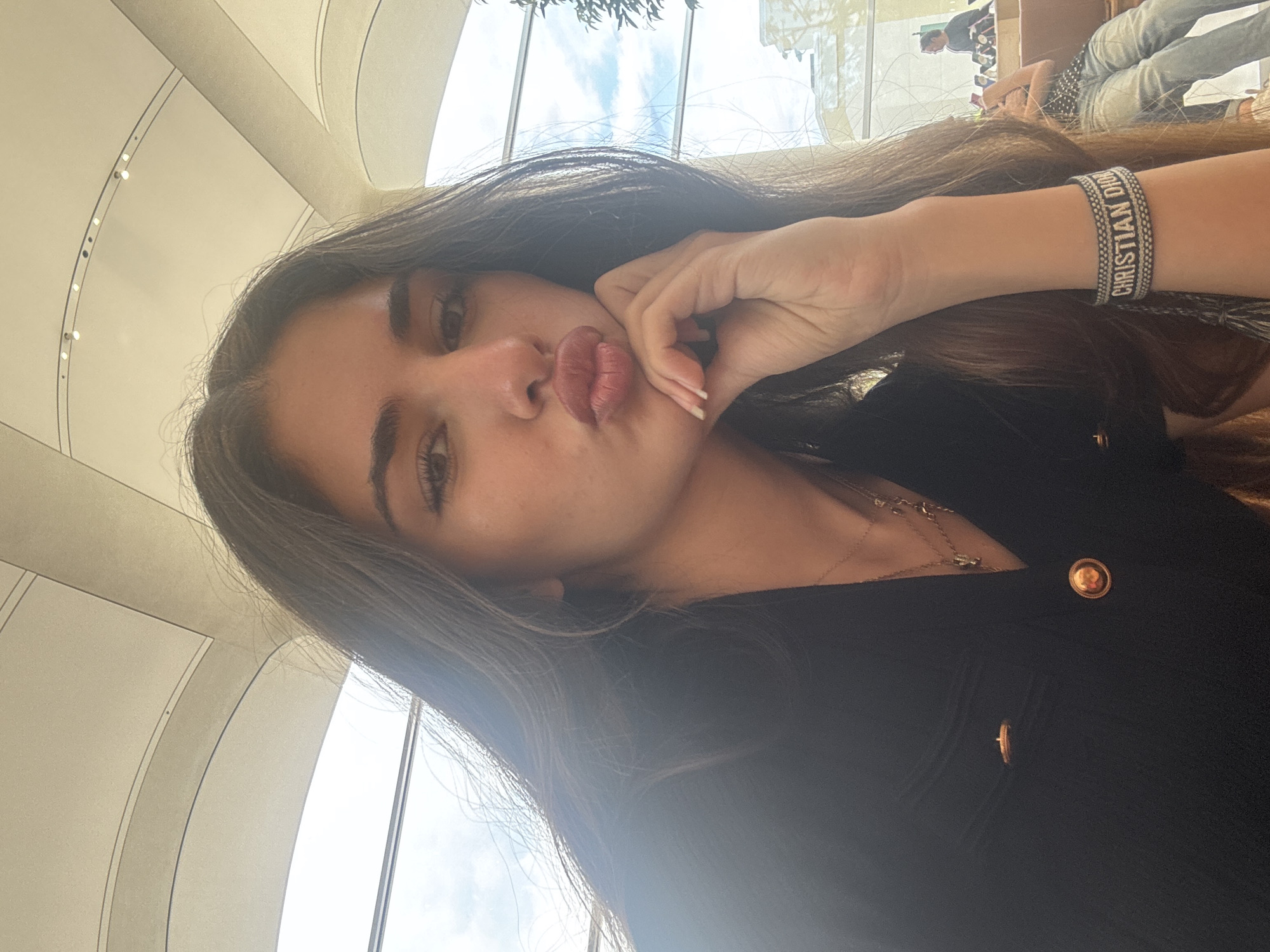 The image is a close-up selfie of a young woman with long, dark hair, puckering her lips and resting her chin on her hand. She is wearing a black top with gold buttons and two necklaces. A bracelet on her wrist reads "CHRISTIAN DIOR." In the background, a glass window shows a blurry view of an outdoor scene with people and buildings, suggesting an urban environment. The ceiling appears to be a curved, modern architectural design, possibly indicating an indoor public space. The image was taken in the city of Aventura, United States of America.