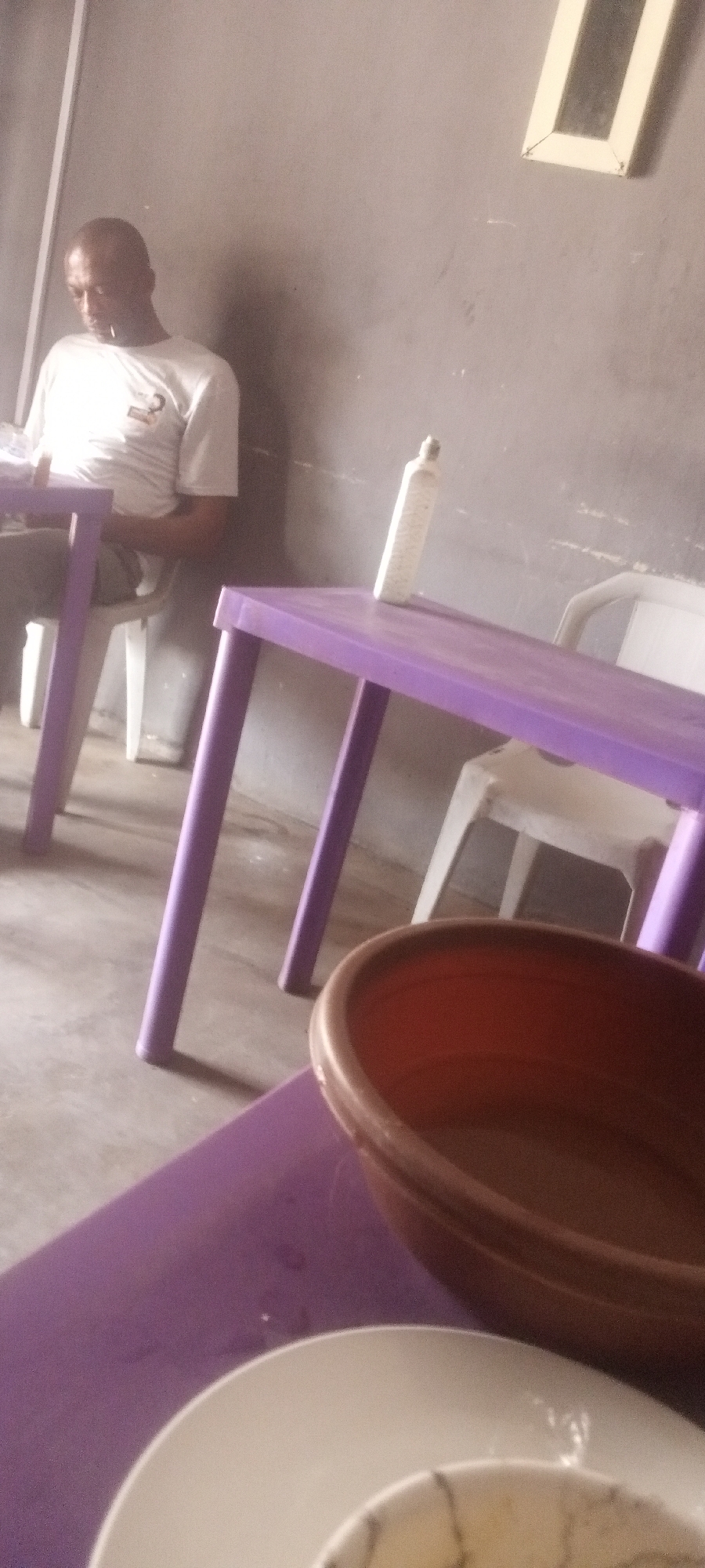 A man is sitting at a table indoors. The table is purple, and there is a white bottle on it. Another table has a bowl and a plate. The man is wearing a white shirt. The walls are gray. A white-framed object, possibly a window or painting, is near the ceiling. The location is Sabon Tasha, Nigeria.