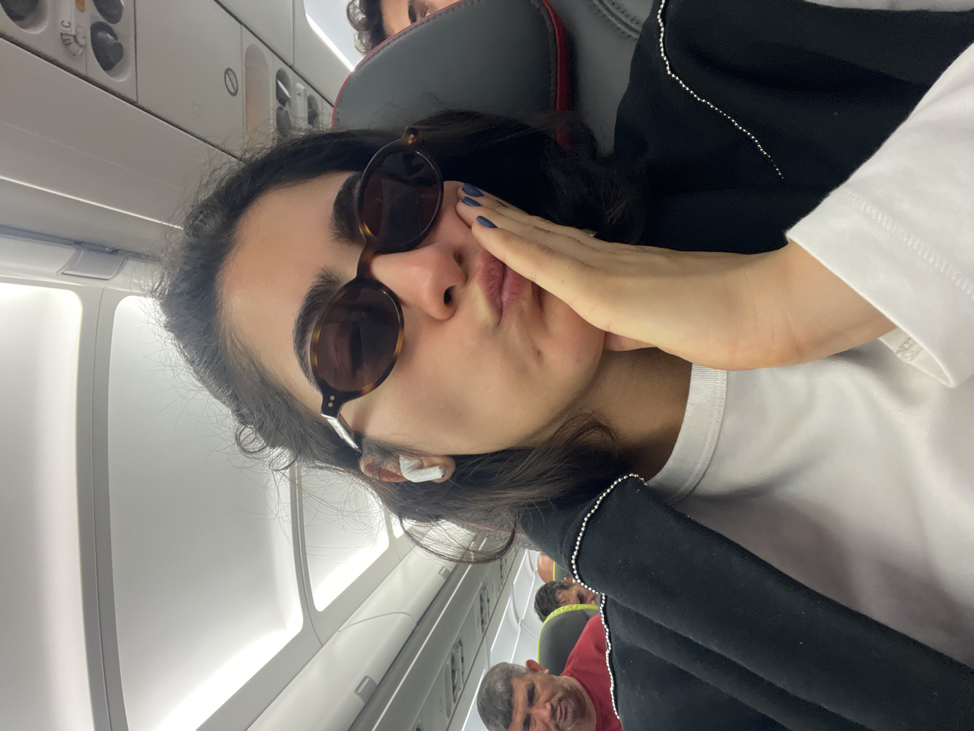 The image shows a close-up selfie of a young woman on an airplane, likely in flight. She is wearing sunglasses, a white t-shirt, and a black jacket adorned with small silver beads. She has her right hand on her cheek and is puckering her lips. White earbuds are visible in her right ear. In the background, other passengers are seated on the plane. One man in a red shirt is visible in the left foreground, and another man is seated behind him. The overhead compartments and lighting of the airplane cabin are also visible. This image was taken inside the cabin of an airplane, not in the city of Moreira, Portugal.