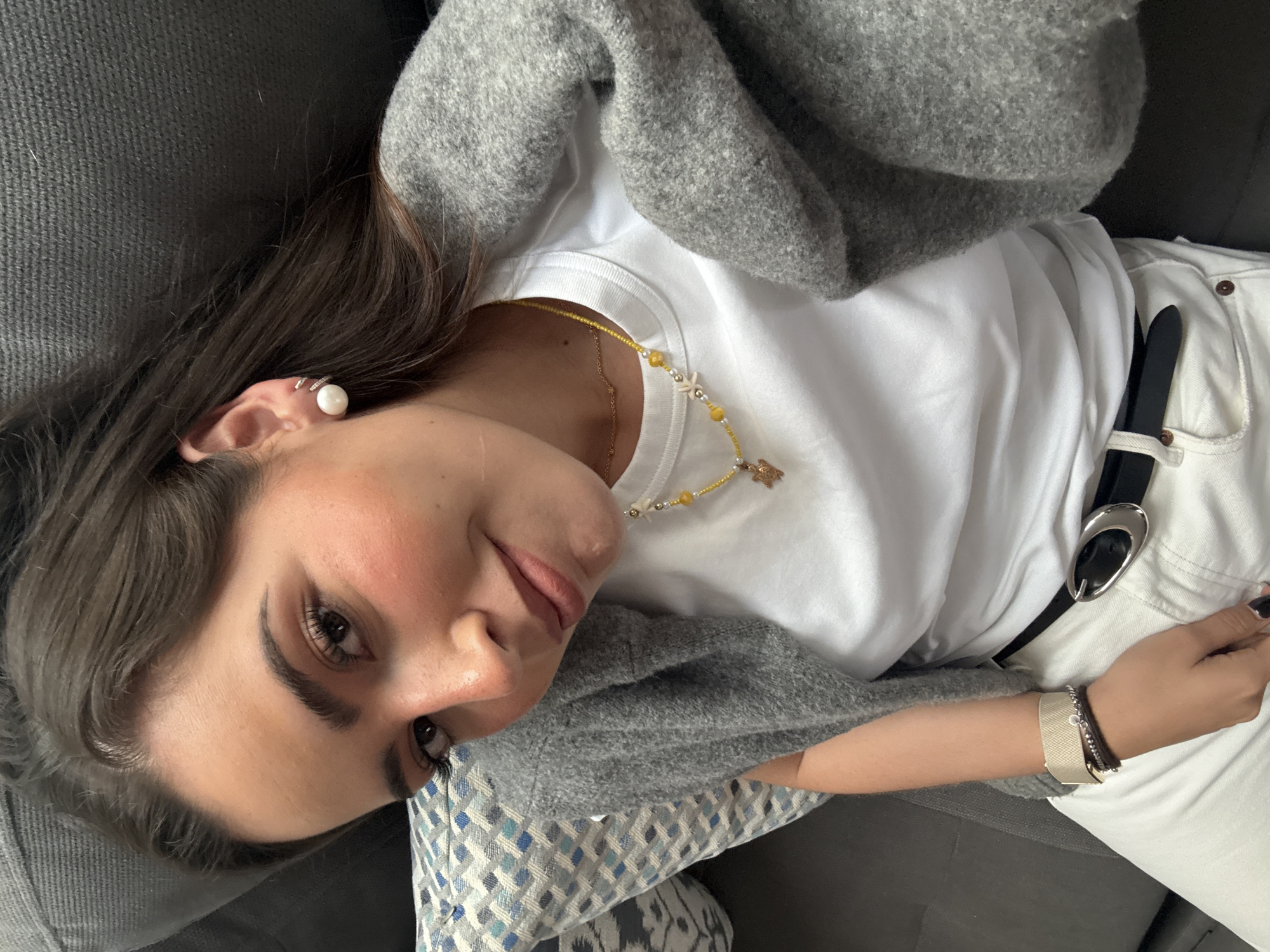 A woman is lying on a gray sofa with a patterned pillow beneath her head. She is wearing a white t-shirt under a gray sweater, along with white pants secured by a black belt with a silver buckle. She is accessorized with a pearl earring, a beaded necklace featuring yellow and white beads, and bracelets on her right wrist. The specific location in San Bernardino Tlaxcalancingo, Mexico cannot be determined from this image.