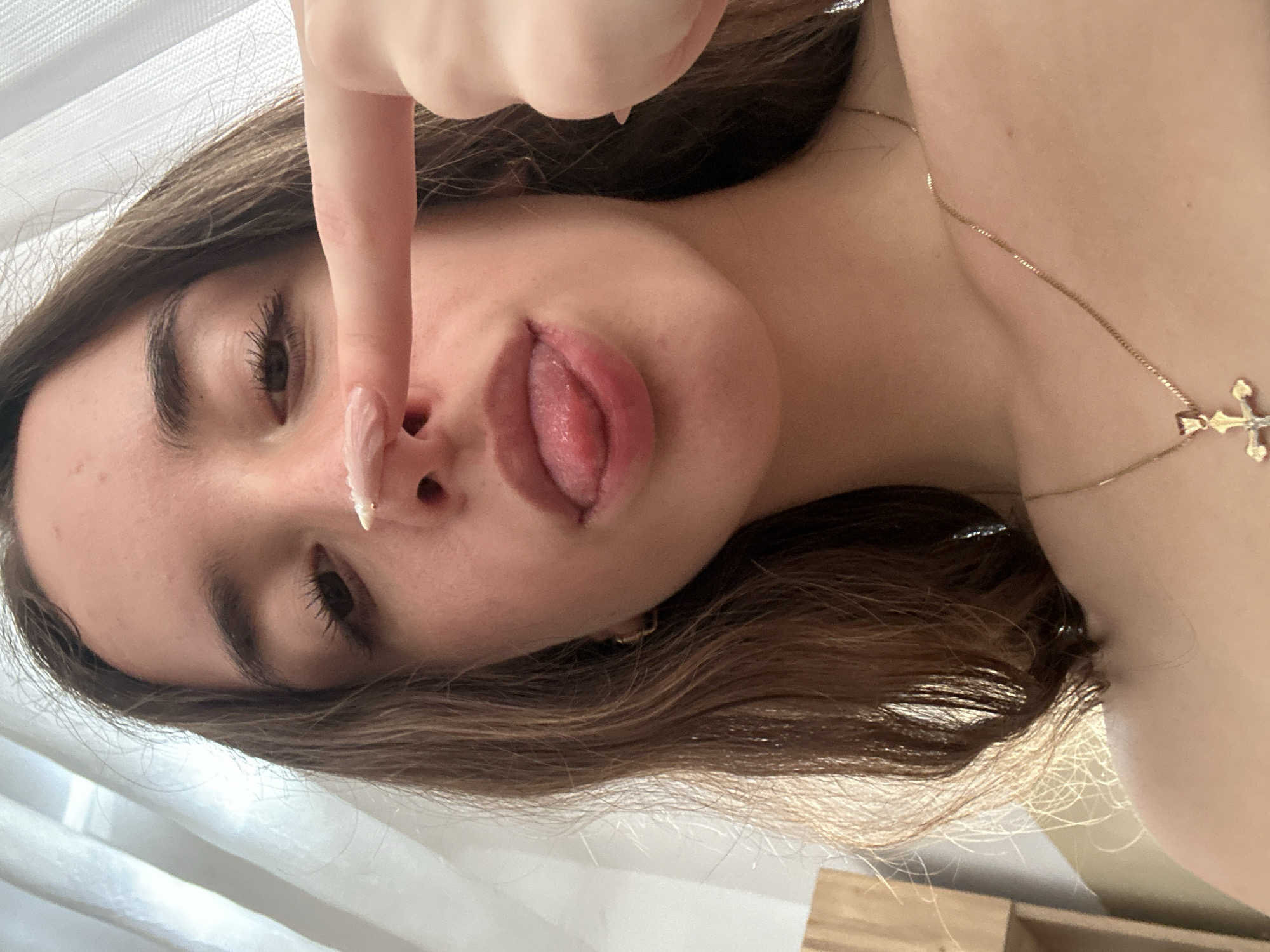A close-up, eye-level, selfie-style photograph shows a young person's face from the nose up, with their tongue sticking out and touching their nose. The person has brown hair, fair skin, and is wearing a gold necklace with a cross pendant. A finger with a light pink nail is positioned to touch the tip of the person's nose. The background consists of sheer, white curtains. No specific location in the city of Shumen, Bulgaria, can be identified from the image.