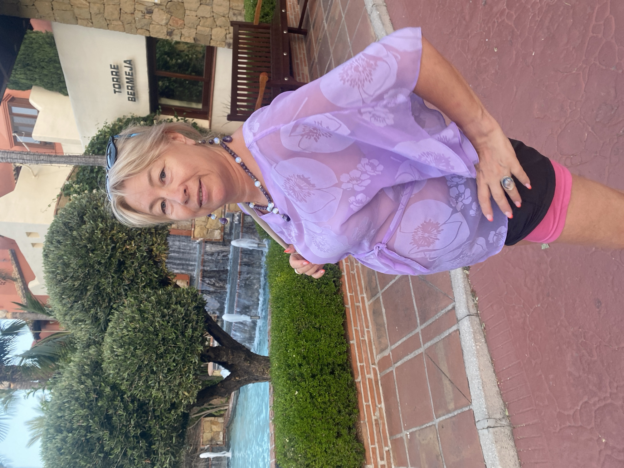 A blonde woman in her late 50s or early 60s is posing for a photo outdoors. She is wearing a sheer lavender cover-up with a floral pattern over a black and pink outfit. She has on a large, ornate ring on her right hand. The woman is smiling and looking towards the camera. Her hair is blonde and shoulder-length, and she is wearing sunglasses on top of her head. A beaded necklace with purple and white beads is around her neck.
The background shows a building with "TORRE BERMEJA" written on it. There is a water feature with fountains and a lush green hedge. The ground is paved with reddish-brown tiles. The lighting suggests it is daytime, likely sunny. The overall setting appears to be a resort or a well-maintained outdoor area, possibly in Estepona, Spain, given the location context. The woman appears happy and relaxed.
