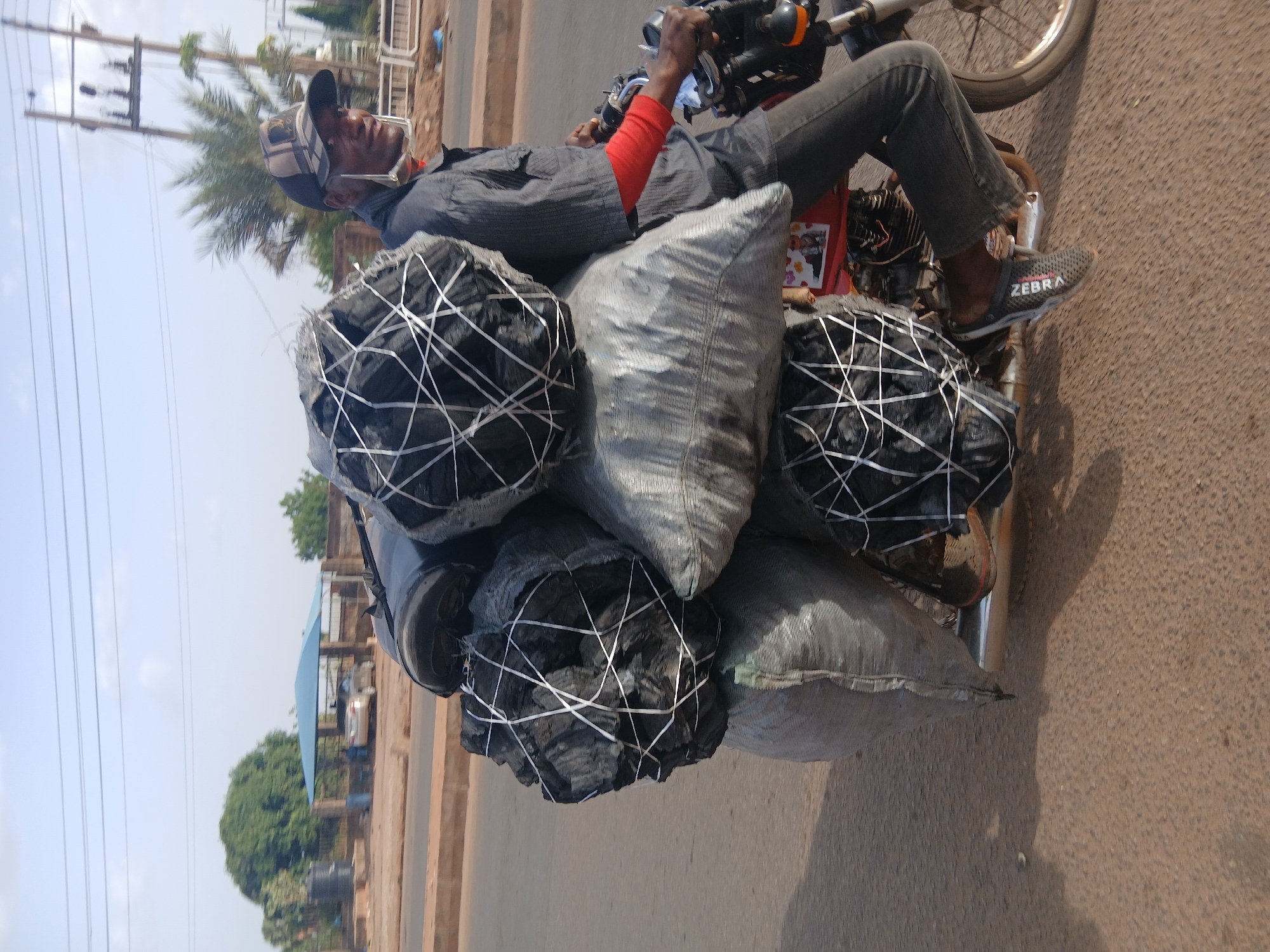 A man is depicted riding a motorcycle heavily laden with goods on what appears to be a paved road in Sabon Tasha, Nigeria. The image captures the man from a slightly elevated side angle as he navigates the road.
**Subjects:** The primary subject is a man, dressed in a dark collared shirt worn over a red long-sleeved garment, dark trousers, and grey and black slip-on shoes with "ZEBRA" visible on the side. He wears a distinctive cap, possibly baseball-style, and a protective face mask or shield. He is seated on a motorcycle, which is obscured by the numerous items being transported. The motorcycle itself is dark-colored, with some red elements visible on the frame. The main cargo consists of multiple large bags, with at least three clearly containing charcoal, bound with white string. Several other grey or silver sacks, likely also containing goods such as charcoal, are stacked around him and on the motorcycle. In the background, there are utility poles, palm trees, and what appears to be part of a building with a light blue roof, hinting at a semi-urban environment.
**Activity:** The man is actively operating the motorcycle, transporting a substantial amount of goods, primarily charcoal. This suggests he is likely a vendor or delivery person engaged in the commerce of fuel.
**Notable details:** The scene unfolds under bright daylight with a clear sky, indicated by the strong illumination and visible shadows. The road surface is a light brown, possibly asphalt or dirt-laced pavement. The sheer volume of goods being carried on the single motorcycle is a prominent feature, highlighting a common method of transportation for bulk items in some regions. The man's attire, including the cap and face mask, suggests protection from sun and dust. The "ZEBRA" text on his shoe is clearly legible.
**Location cues:** The presence of palm trees, utility infrastructure, and the general appearance of the road and background buildings are consistent with the provided context of Sabon Tasha, Nigeria.