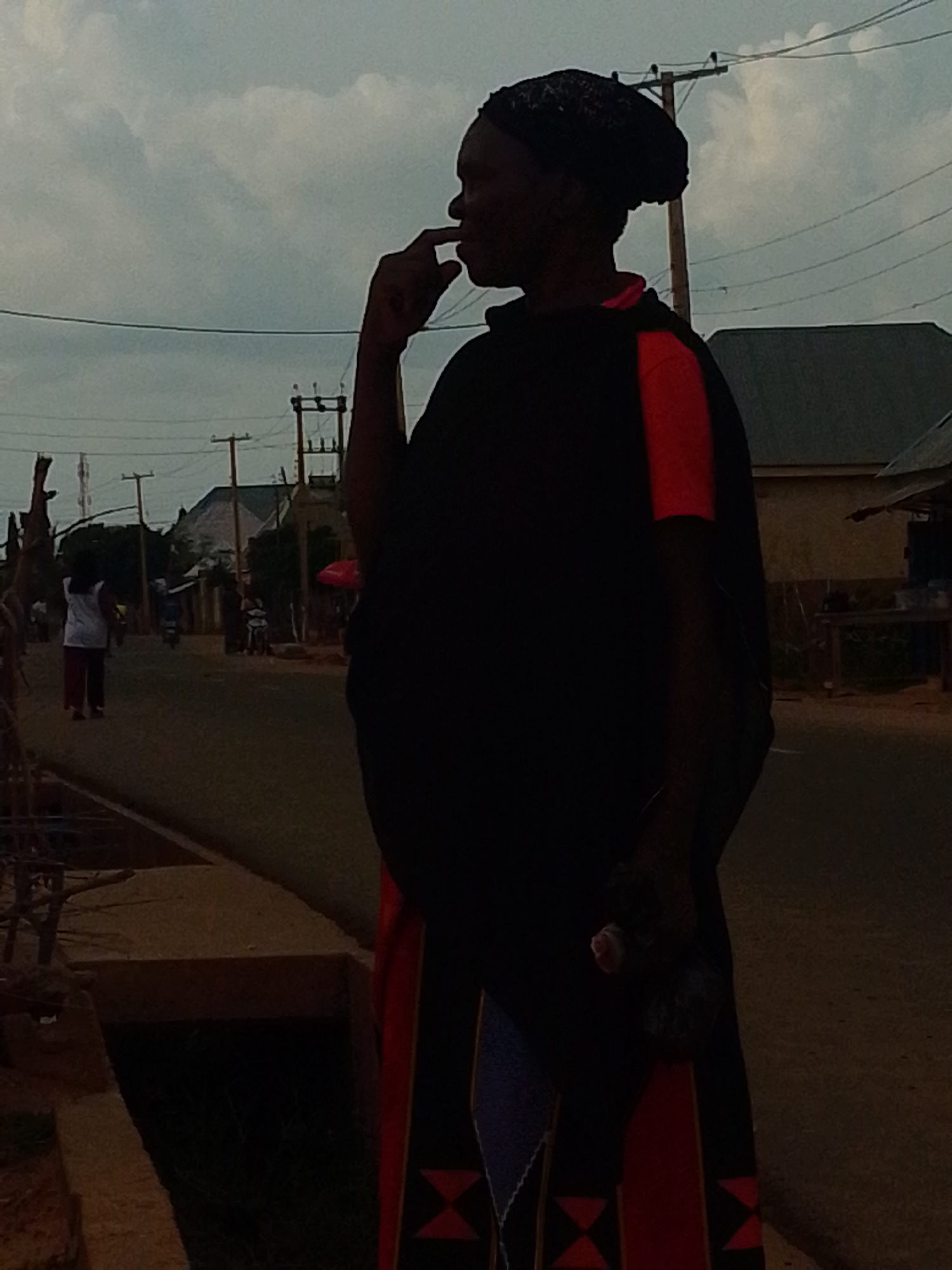 A person stands in silhouette against a cloudy sky, facing right with their hand near their mouth. They wear a dark head covering and a dark garment with red accents on the sleeves and hem. In the background, a street scene unfolds in Tempure, Nigeria. The street is lined with buildings and utility poles with visible wires. Several people are walking and riding motorcycles in the distance. The overall lighting suggests it is either early morning or late evening.