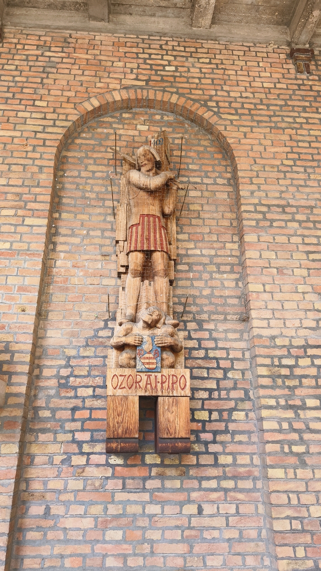The image shows a wooden sculpture mounted on a brick wall in Szeged, Hungary. The sculpture depicts a standing male figure with a headdress, a red and white striped skirt, and he is holding two long spears. Below him, a smaller carved figure is holding a shield with a coat of arms. A wooden plaque beneath this reads "OZORAI-PIPO" in red lettering. The sculpture is set within a brick archway, and the surrounding wall is constructed of reddish-brown bricks with gray mortar. The overall scene is outdoors, with a concrete beam visible at the top of the frame.