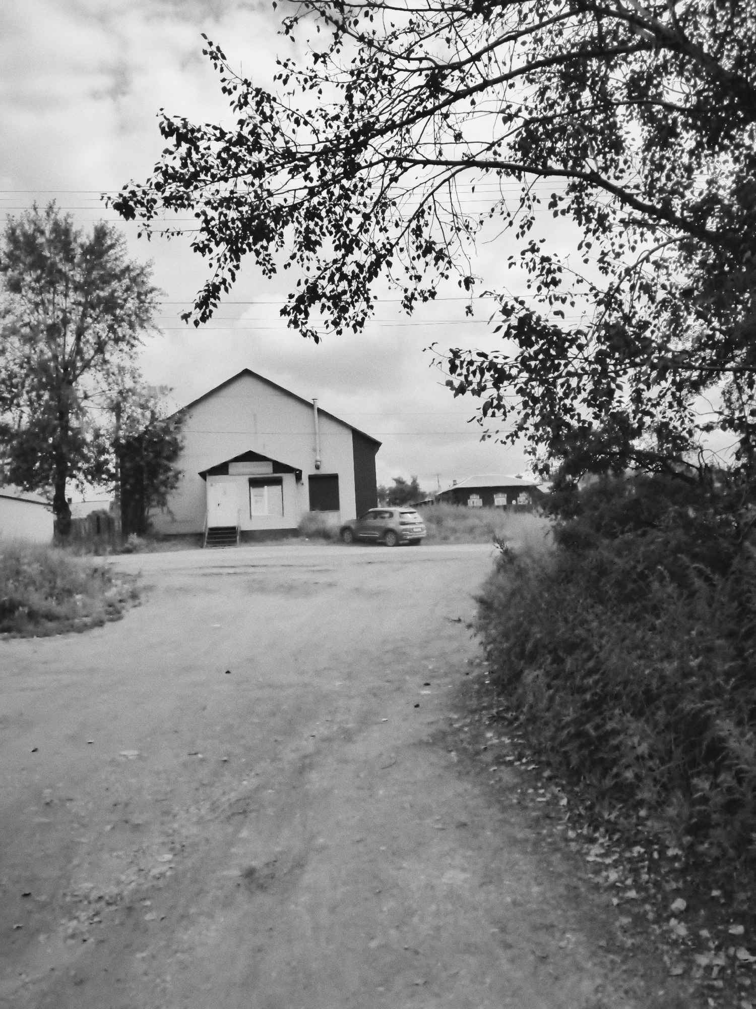 The black and white photograph captures an outdoor scene in Cheremkhovo, Russia. In the foreground, a dirt road curves towards the right. The left side of the road is bordered by patchy vegetation and a leafless tree. Dominating the midground is a large, rectangular building with a gabled roof and a dark, rectangular window. A small, covered entrance with stairs leads to a door. A silver car is parked to the right of the building, facing away from the viewer. Further in the background, another building with multiple windows is visible behind a thick line of foliage. The sky above is overcast with scattered clouds, and branches of a leafy tree hang over the top of the image. The scene suggests a rural or semi-urban setting.