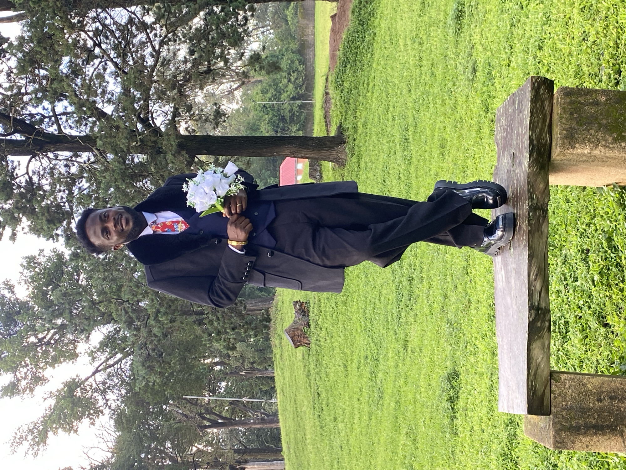 A Black man, appearing to be in his late 20s to early 30s, is standing on a stone bench in a park. He is wearing a dark suit with a navy vest and a black overcoat with a fur collar. He has on a red and white patterned tie and a gold bracelet on his left wrist. In his right hand, he holds a bouquet of white flowers. His left leg is crossed over his right, and he is looking towards the camera with a slight smile. The background consists of a lush green grassy field with scattered trees and a red-roofed building in the distance. The location is identified as a park in the city of Jos, Nigeria.
