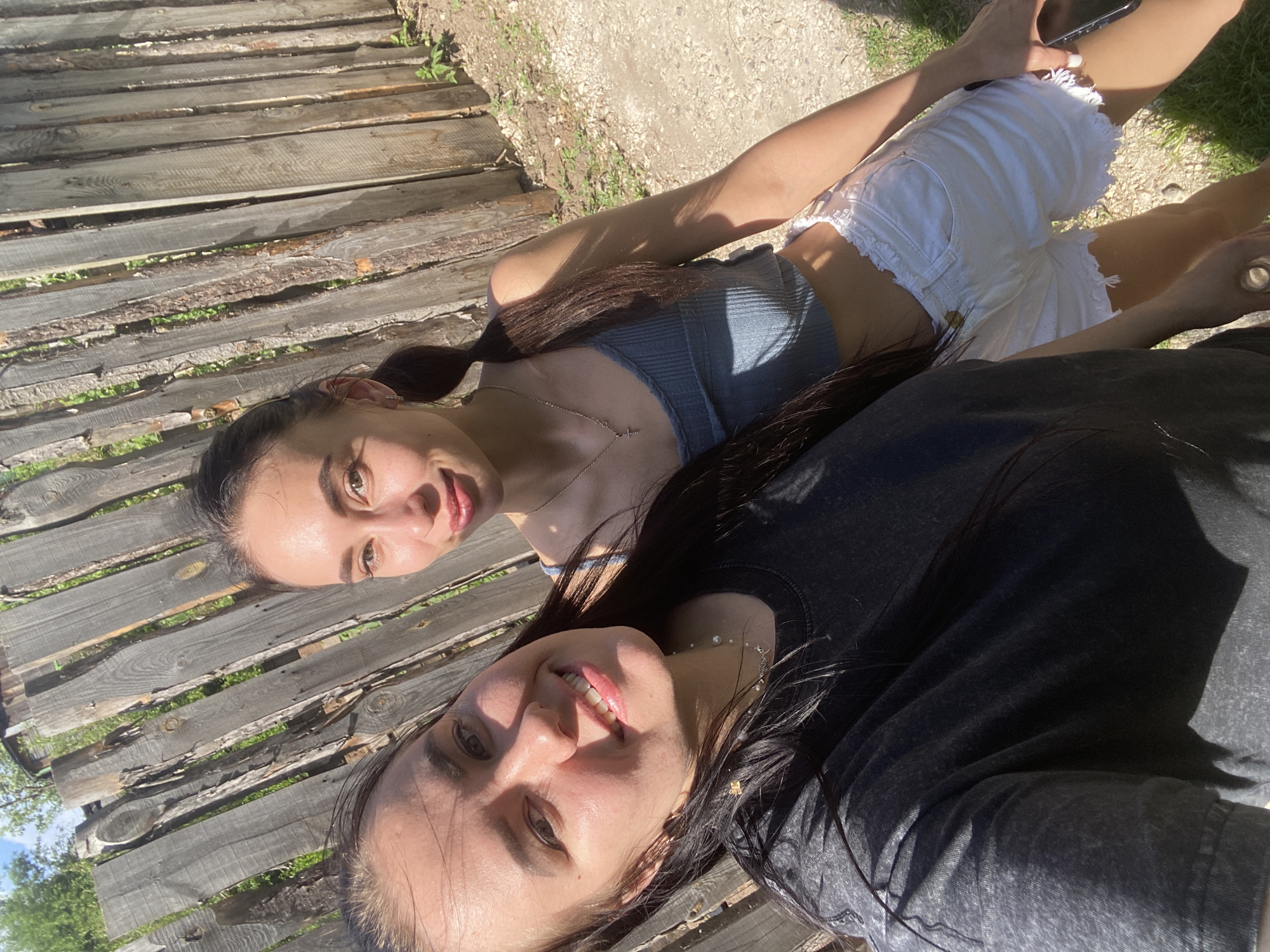 Two young women are posing for a selfie outdoors. The woman on the top wears a gray tank top and white shorts, while the woman on the bottom wears a dark gray t-shirt. They are sitting on a wooden deck in a sunlit area. A mobile phone is visible in the hand of the woman on top. The location is likely Chaikovsky, Russia, based on image details.