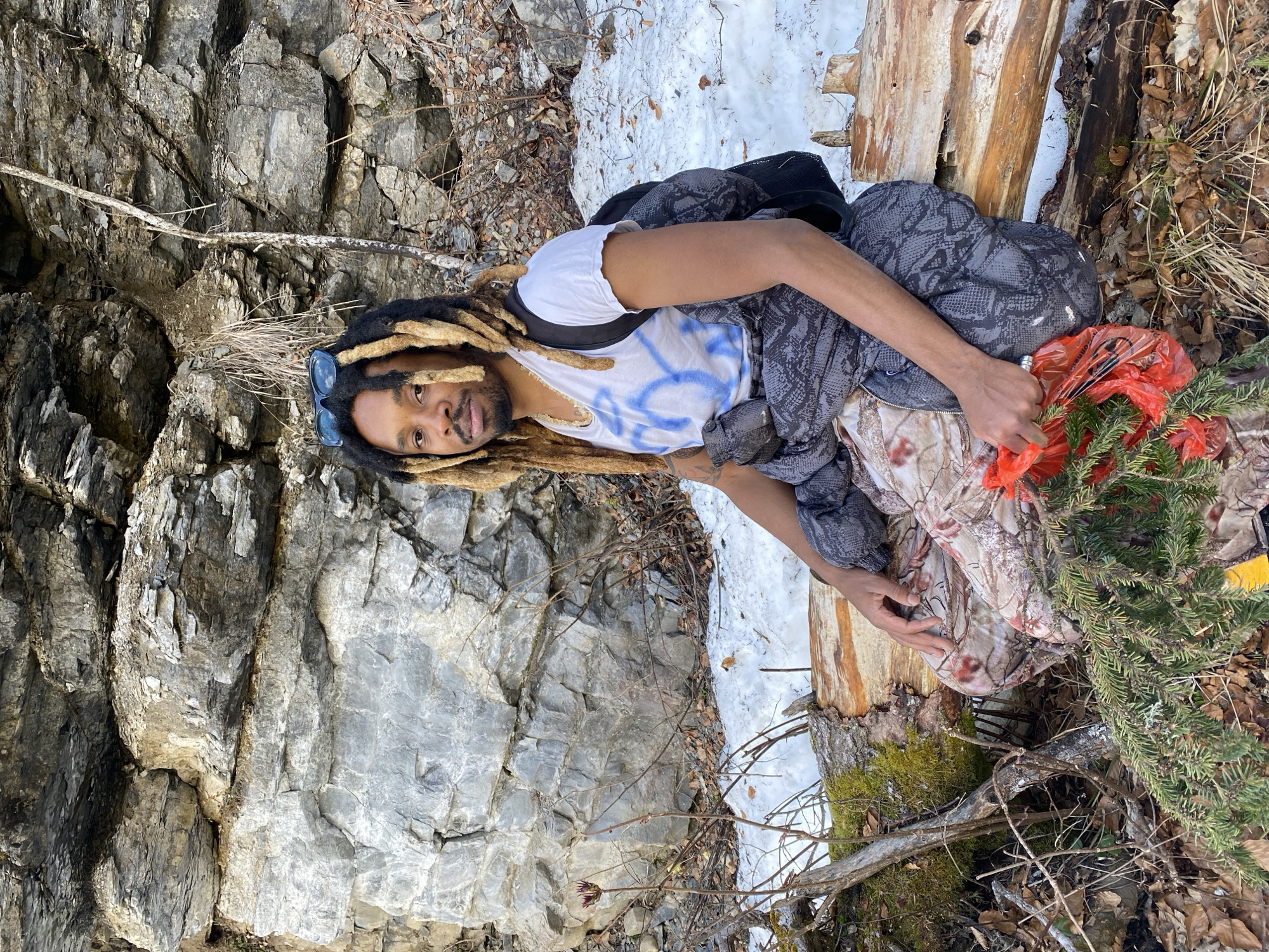 A person is sitting outdoors on a log or fallen tree trunk amidst a mix of natural elements. The person has dreadlocks, sunglasses on their head, and is wearing a white t-shirt with blue markings and a gray jacket. They are holding a red plastic bag filled with green pine branches. There's snow on the ground, and dead leaves scattered around. To the left of the person is a large rock face.
The location, Saint-Michel-les-Portes, France, cannot be verified from the image alone.