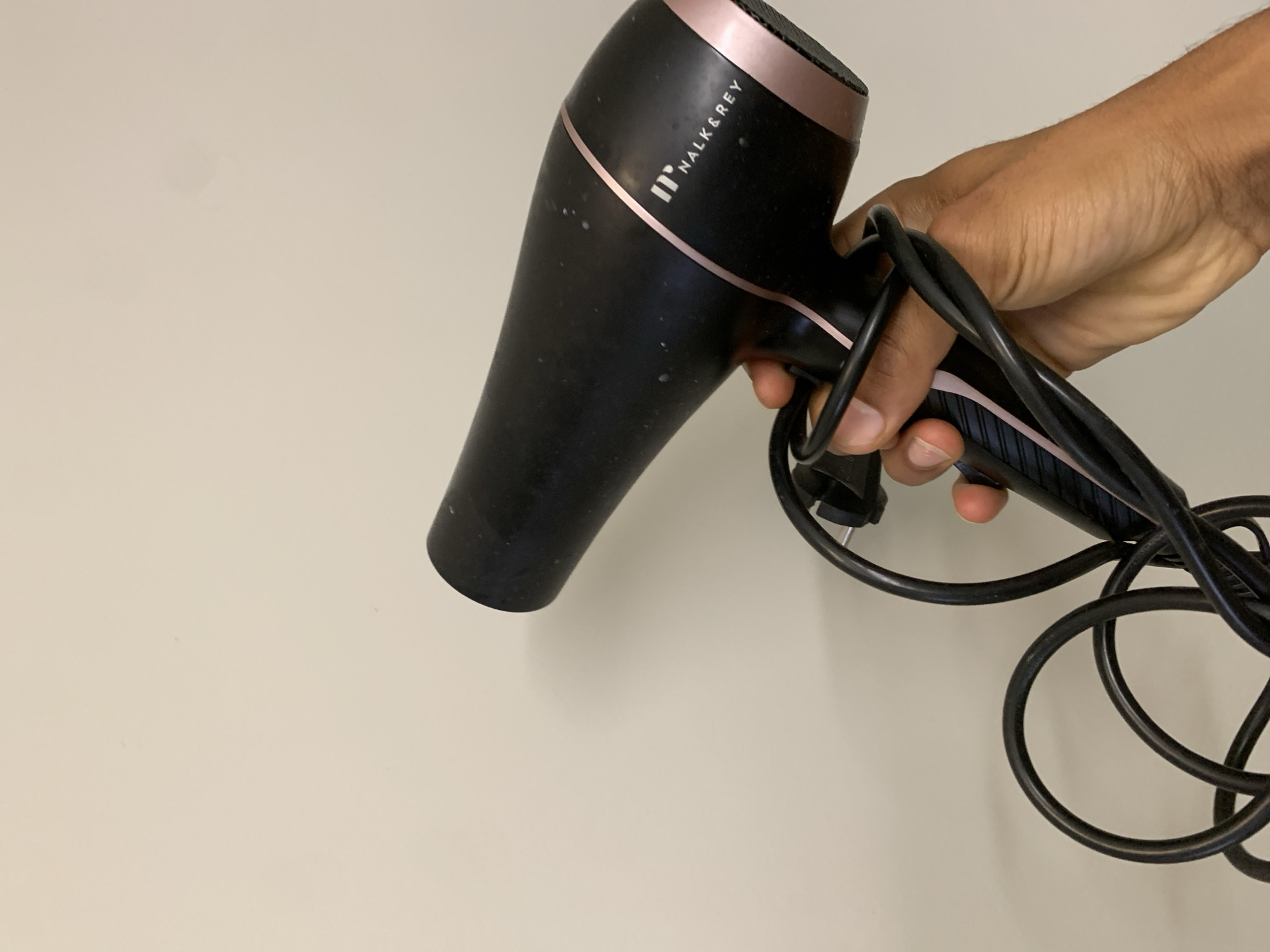 A black hairdryer with rose gold accents is held in a person's hand against a plain, light-colored wall. The hairdryer has the brand name "NALK & REY" printed in white. The cord of the hairdryer is coiled around the handle and dangles below. The background appears to be an indoor wall, with no discernible architectural features or context to indicate a specific location in Barcelona, Spain.