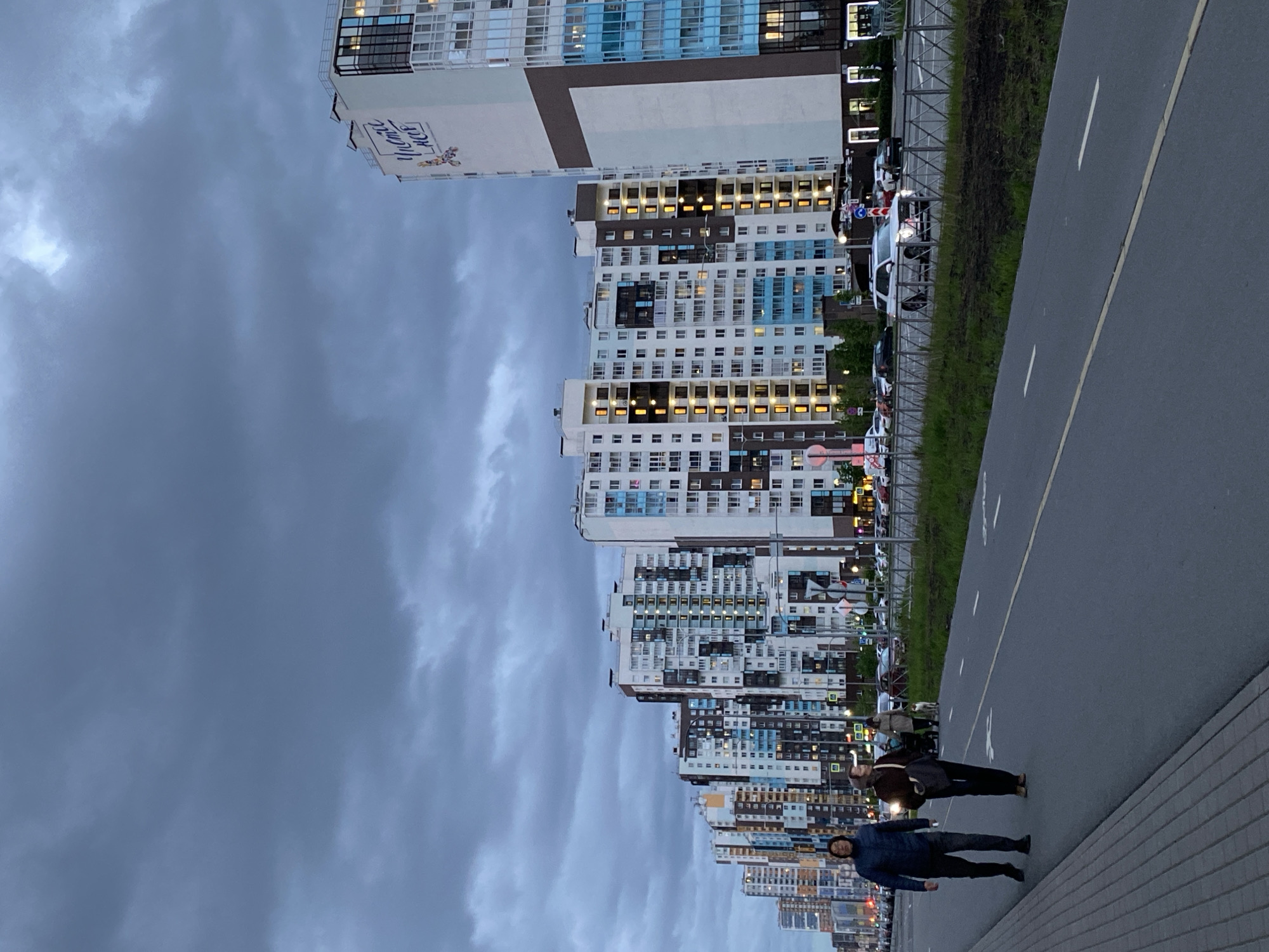 The image shows a street scene in Saint Petersburg, Russia, with high-rise apartment buildings lining the left side of the frame. The buildings are of modern architecture, with varying color schemes and numerous windows, many of which are illuminated, indicating nighttime. A wide paved path, likely a sidewalk or bike path, runs along the right side of the image, separated from the buildings by a thin strip of vegetation and a metal fence. Two individuals are walking on the path. The first person is wearing a dark blue jacket and dark pants and is slightly ahead of the second person. The second person is wearing a brown jacket, dark pants, and carrying a bag. The sky above is cloudy and overcast, contributing to the scene's dim lighting. Cars are parked along the street in front of the apartment buildings.
