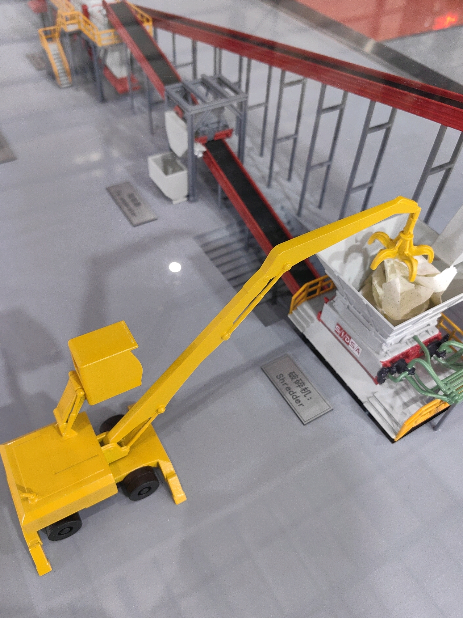 The image depicts a miniature model of an industrial waste processing facility. The model includes conveyor belts, processing machinery labeled "Shredder," and a yellow crane-like vehicle. The facility appears to be a scale model, possibly for display or educational purposes. There are no people visible in the image. The environment is a flat, grey surface, representing the ground. The lighting suggests an indoor setting. The specific location of this model within the city of Mumbai, India, cannot be determined from the image alone.