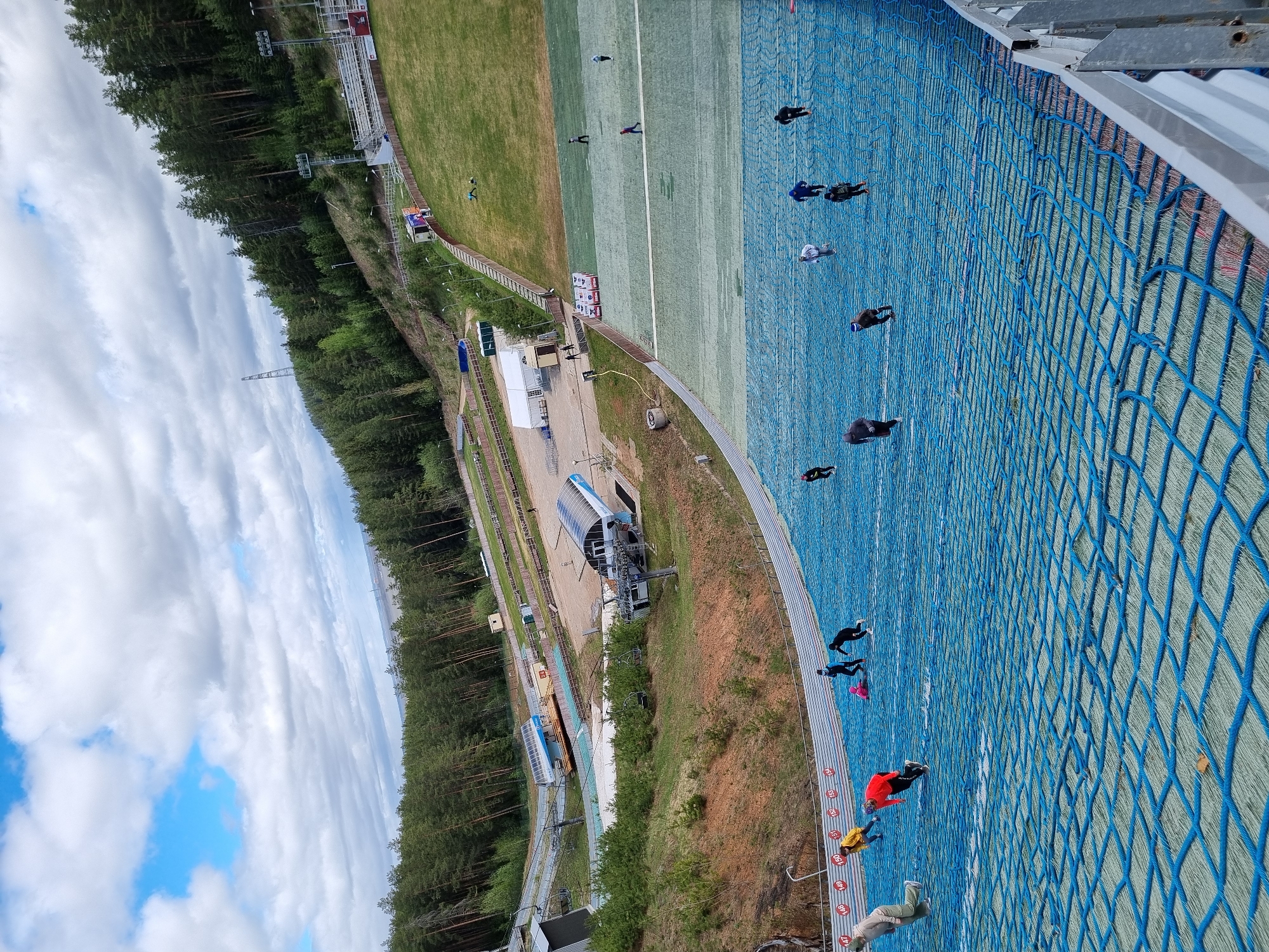 The image shows a ski jumping hill in Прикамский, Russia. The synthetic surface of the hill is visible, and several people are walking on it. A blue net is partially obstructing the view. In the background, there is a ski lift and a forest. The sky is cloudy.