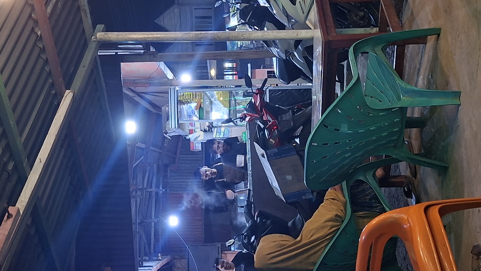 The image depicts an outdoor dining area at night in Keudegeudong, Indonesia, possibly a warung or street food stall. In the background, two men are working behind a counter. One man, wearing a brown apron, is actively cooking, with steam rising from his workstation. Another man to his right, wearing a black t-shirt, appears to be assisting or serving. A third man, partially visible on the far left, is also looking towards the counter.
In the foreground, several plastic chairs are arranged around tables. Two green chairs are prominent, one occupied by a person wearing a yellow hijab and a patterned skirt. An orange plastic chair is stacked in front of them. Motorcycles are parked behind the counter area, with one red and one white motorcycle clearly visible. The environment is lit by artificial lights, casting shadows and creating a warm, nocturnal ambiance. The roof structure above is made of corrugated metal and wooden beams.