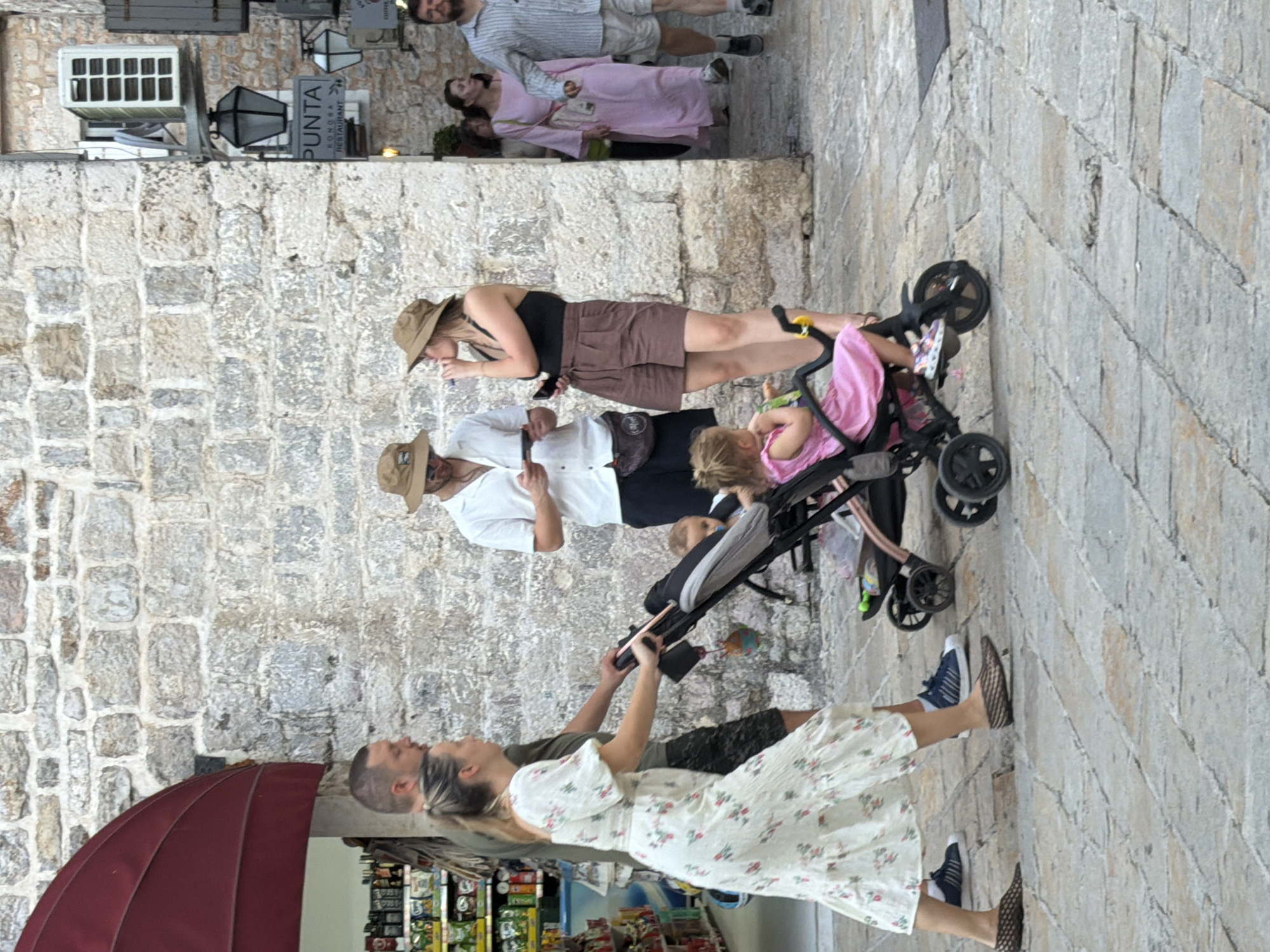 In Budva, Montenegro, a family is captured in a bustling street scene. A woman in a white floral dress pushes a stroller containing a young child dressed in pink. Beside her, a man in a green shirt walks with his arm around her. In the background, another couple stands near a stone wall, the woman in a tan hat and black top, and the man in a white shirt and tan hat, both looking at their phones. Further back, several other individuals are walking through a passageway. The environment is characterized by old stone buildings and a paved street, suggesting a historic part of the city.