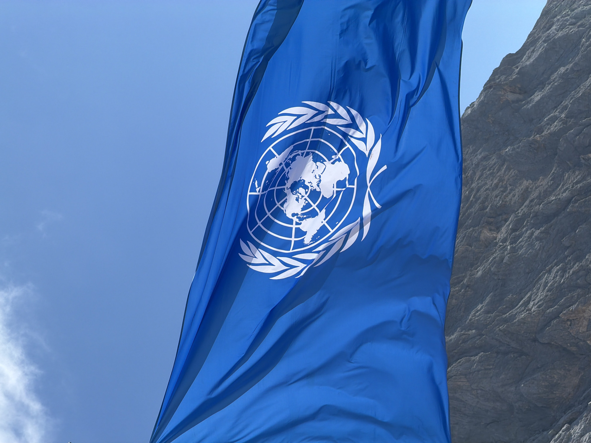 A blue flag with the United Nations emblem in white is prominently displayed, waving against a clear blue sky with scattered clouds. The UN emblem, featuring a globe surrounded by olive branches, is clearly visible on the flag. Below the flag, the rugged, grey surface of a mountain range is visible. The image is a vertical shot. The provided hints do not suggest a specific location in Garmisch-Partenkirchen, Germany.