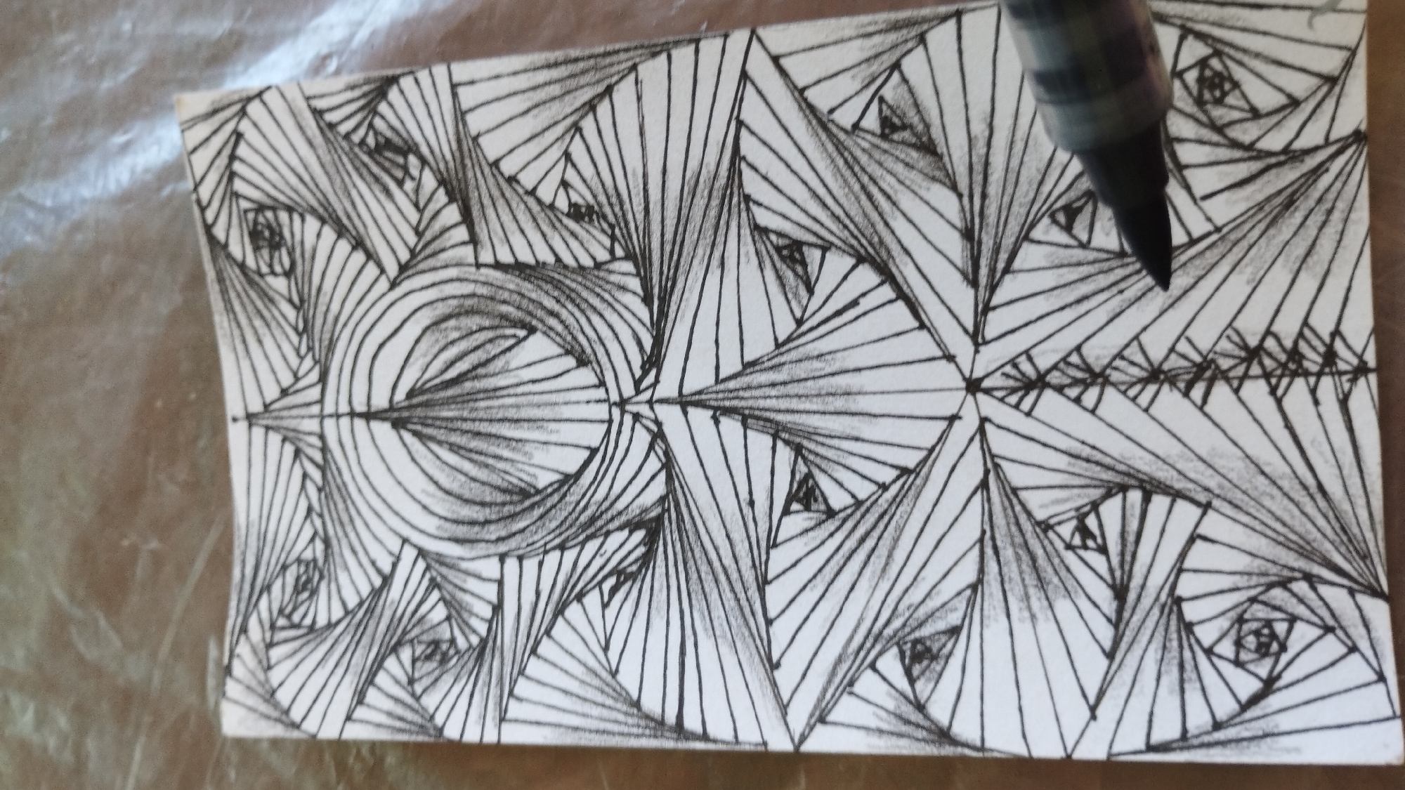 The image shows a close-up of a piece of white paper with a black ink drawing. The drawing features a complex geometric pattern created with thin, parallel lines that form spiraling shapes and triangles. The pattern is abstract and does not depict any recognizable objects, people, or scenes. The lines create a sense of depth and dimension, with some areas appearing darker due to the density of the lines. In the top right corner of the image, the tip of a black marker pen is visible, suggesting that the drawing is in progress. The paper is placed on a textured surface, possibly a mat or a piece of fabric, which is out of focus and appears to be a neutral, muted color. The lighting is even, with no strong shadows, and the overall impression is of a focus on the artistic creation. There is no visible text in the image.