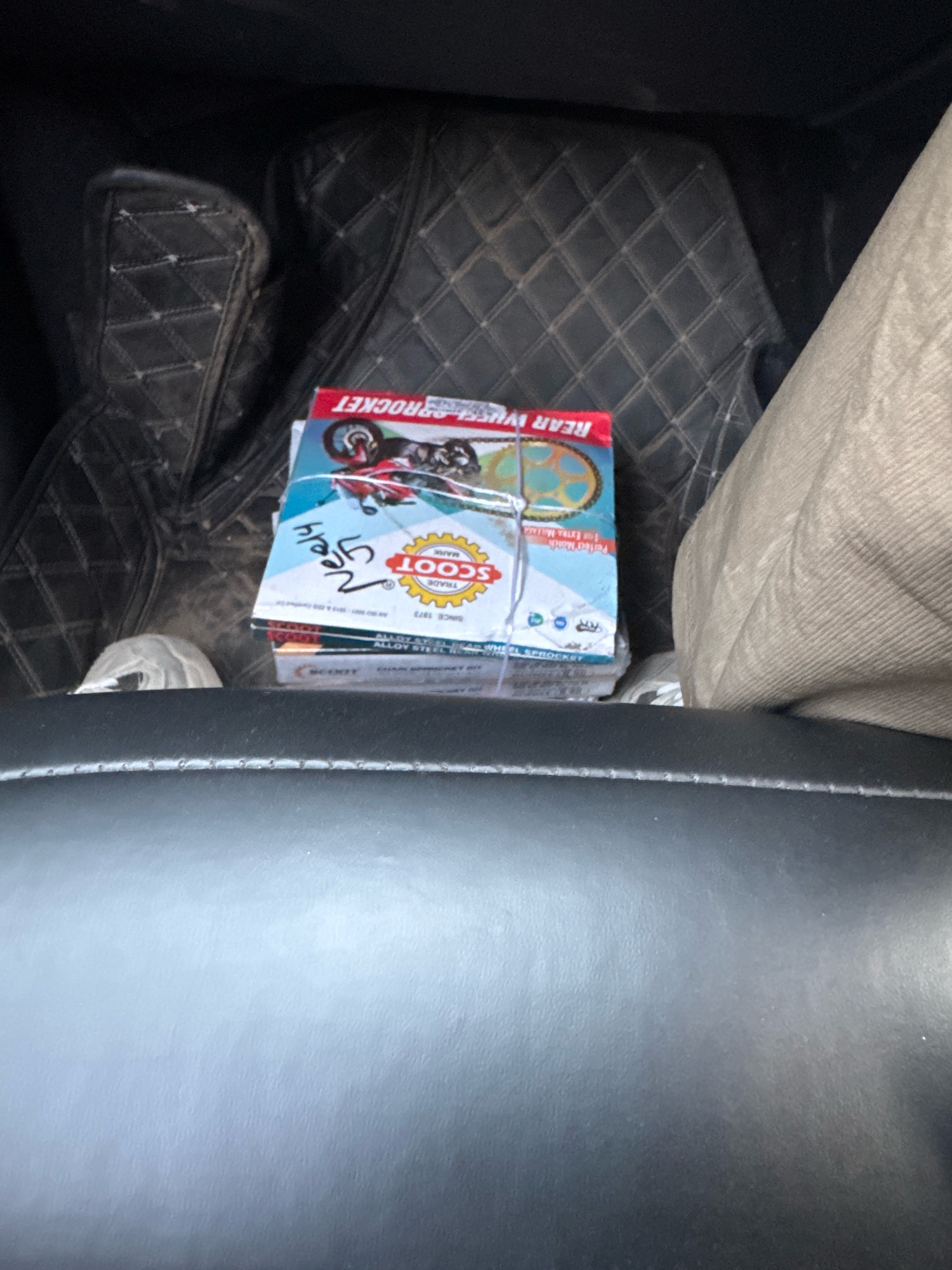 The image shows a stack of several boxed motorcycle sprockets placed on the floor mat inside a vehicle. The boxes have "SCOOT" and "REAR WHEEL SPROCKET" branding. A red and blue graphic of a motorcycle is visible on the top box. A handwritten label "JAIS 25/5/24" is present on the top box. The vehicle's interior, including black diamond-stitched floor mats and a person's leg in light-colored pants, is also visible. The scene appears to be in the city of Ladwa, India, based on the provided context.
