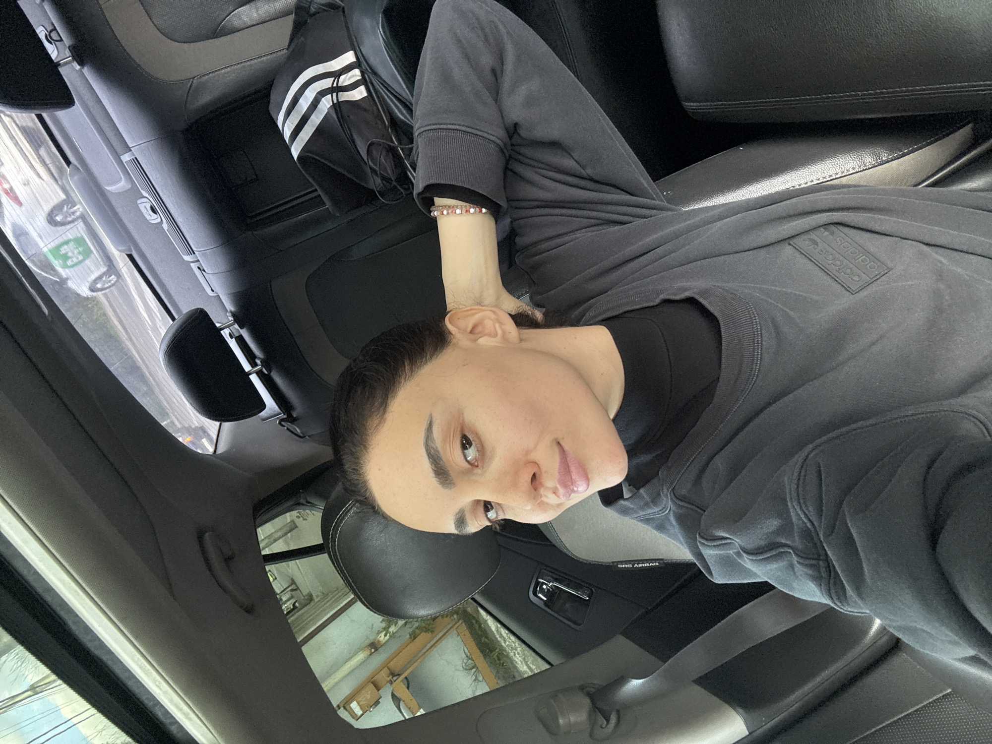 A person is inside a vehicle. The location is suggested to be in Tbilisi, Georgia, based on the presence of green-colored delivery vehicles seen through the car window. The person is wearing a gray sweatshirt with the Adidas logo and a black turtleneck underneath. The person is fair-skinned with dark hair slicked back and is wearing a bead bracelet on their left wrist. An Adidas bag is on the back seat. The interior of the vehicle is gray and black.
