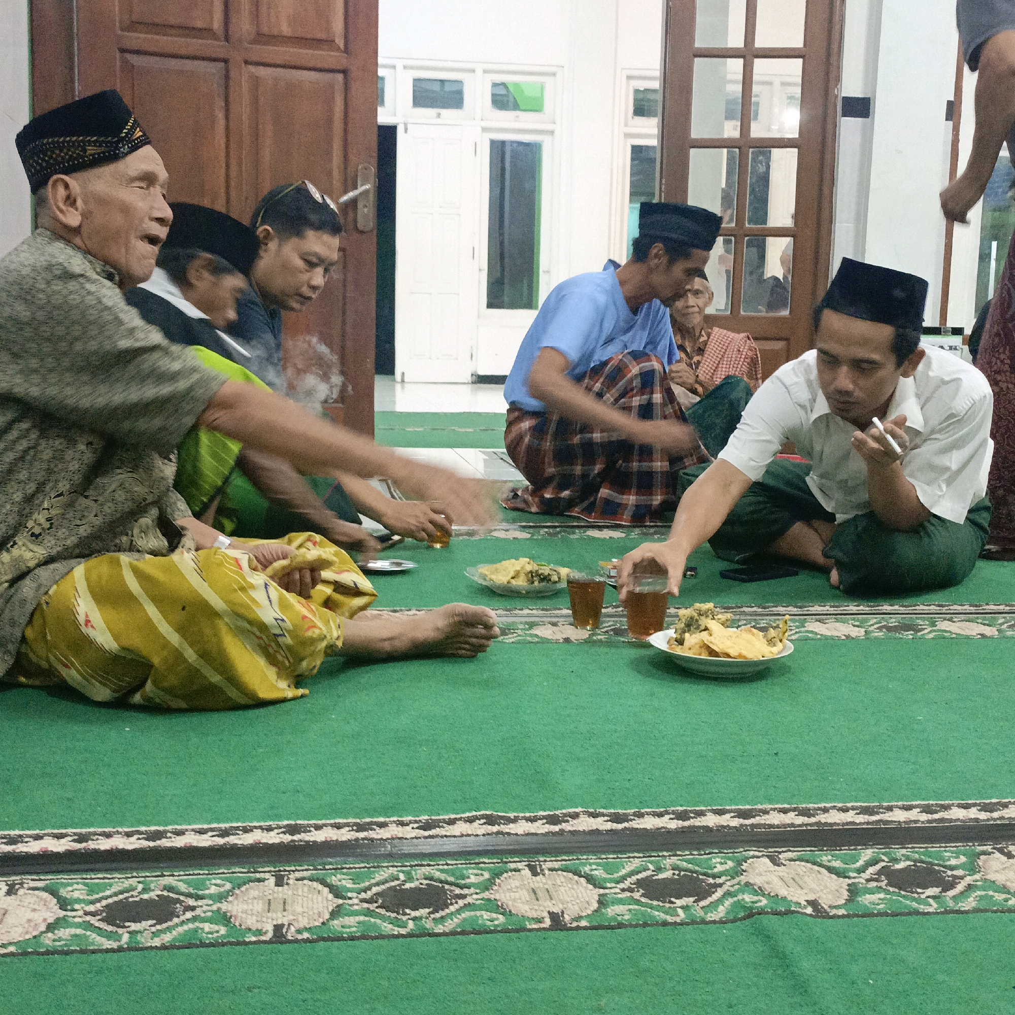 The image depicts a group of men gathered indoors, seated on a green carpeted floor. The room appears to be a home or a community gathering space, possibly a mosque or similar religious building, given the attire of the men and the presence of a carpet. The background features wooden doors and windows. Several men are wearing traditional peci hats. In the foreground, some men are seen interacting with food and drinks, with plates of fried snacks and glasses of tea on the floor. One man is smoking. The overall atmosphere suggests a casual social gathering. The image does not contain enough information to definitively identify the specific location in Salatiga, Indonesia, but the cultural context points to a scene that could commonly occur in such a city.