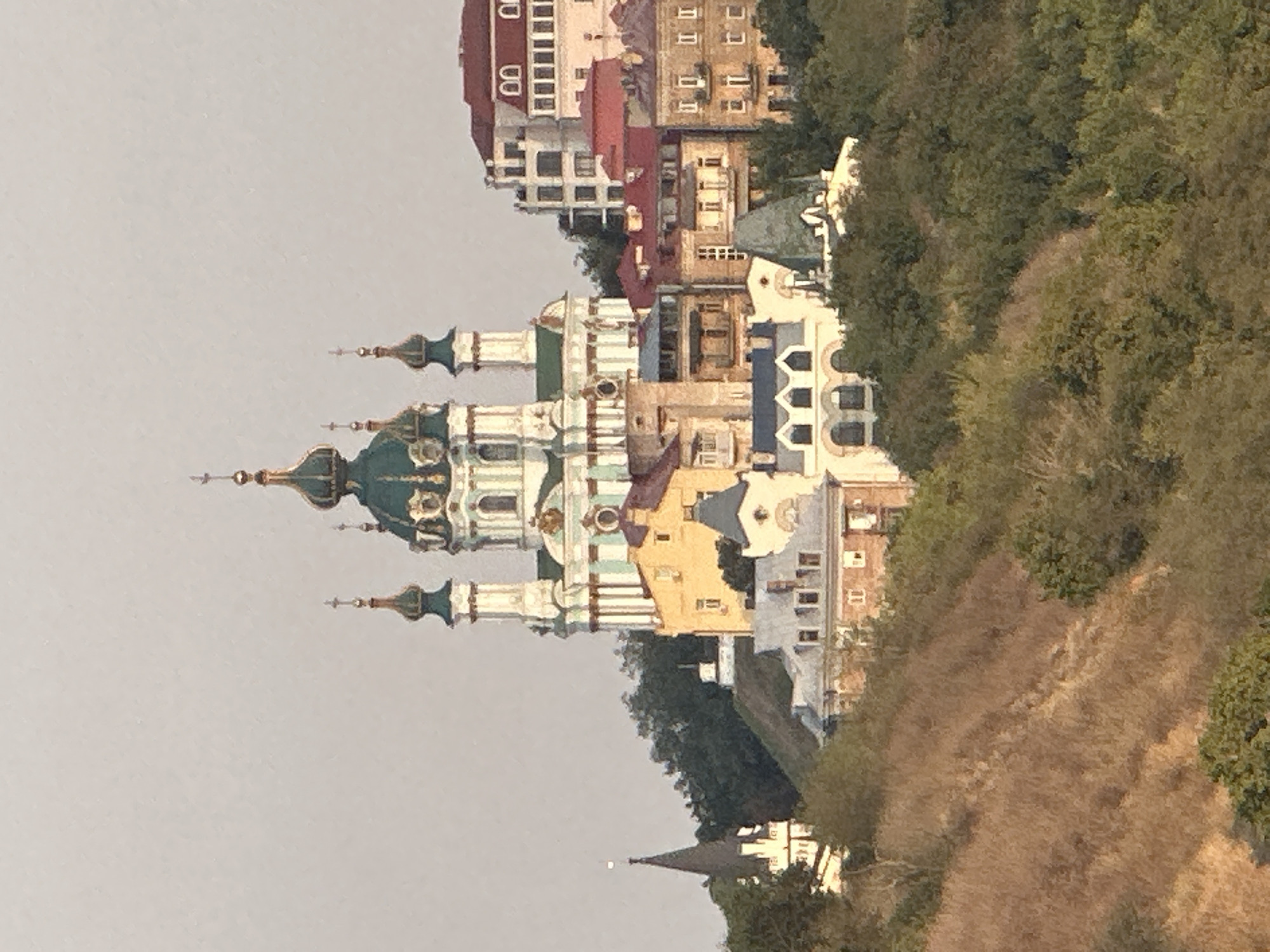 The image depicts a view of Kyiv, Ukraine, focusing on Saint Andrew's Church, an iconic baroque church perched atop a hill. The church features distinctive green domes topped with crosses and a white and light blue facade. Surrounding the church are a cluster of other buildings with red and grey roofs, typical of the Podil district. The scene is set against a backdrop of lush green trees and a gently sloping hill covered in dry grass in the foreground. The sky is a pale, washed-out color, suggesting either early morning or late afternoon light. No people are visible in the image. The overall impression is a serene, picturesque cityscape.