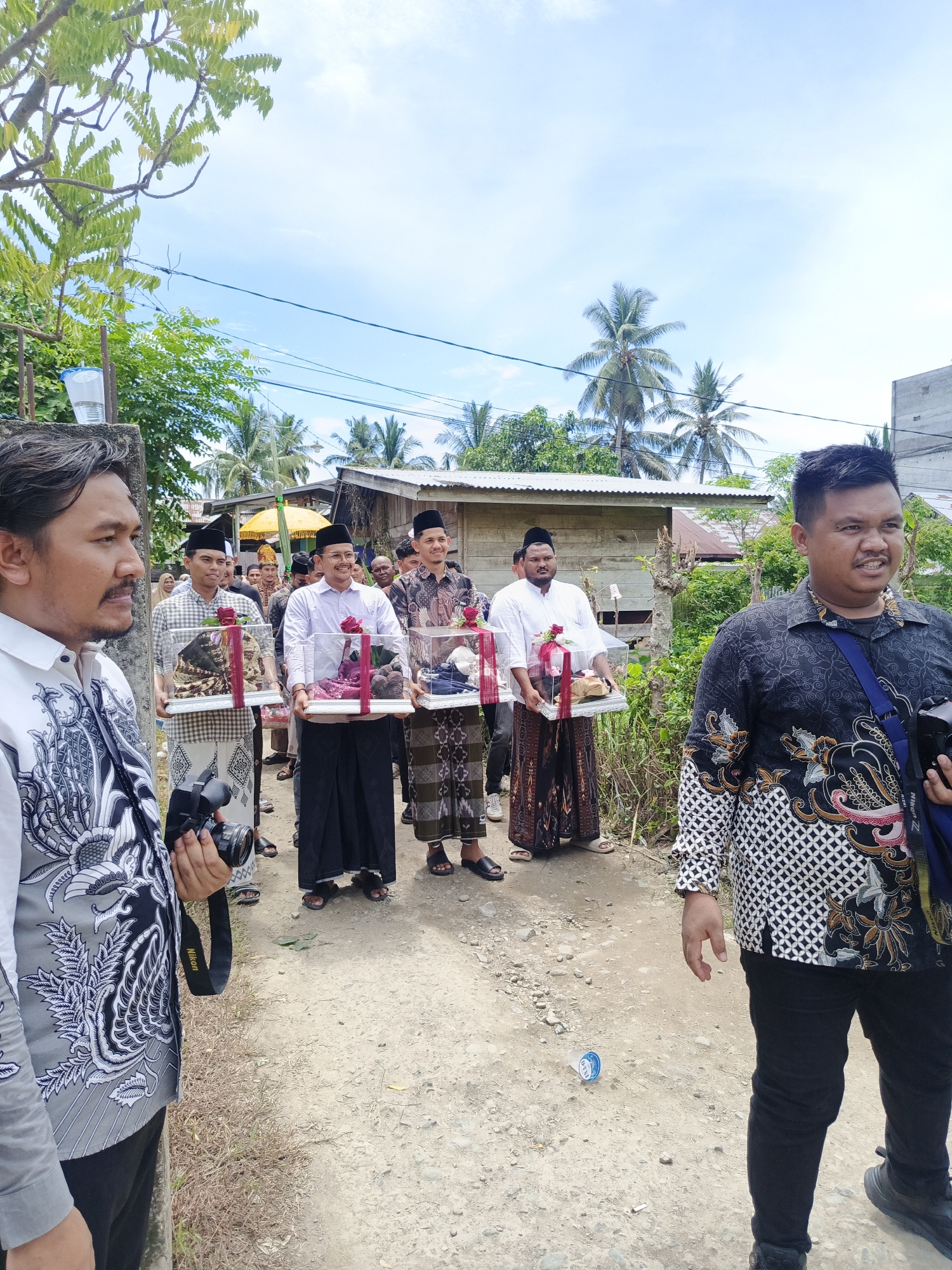 In Meuliek, Indonesia, a group of men are walking in a procession on a dirt path, carrying gifts in clear plastic boxes. The men are dressed in traditional attire, including sarongs and peci hats. The gifts appear to be various items, possibly for a wedding or other ceremony, wrapped in red ribbon and some decorated with red flowers. The environment is outdoors with a background of palm trees and simple buildings. The sky is partly cloudy. One man in the foreground on the left is holding a camera, and another man on the right is also holding a camera bag strap. The image captures a moment of tradition and community event in a rural Indonesian setting.