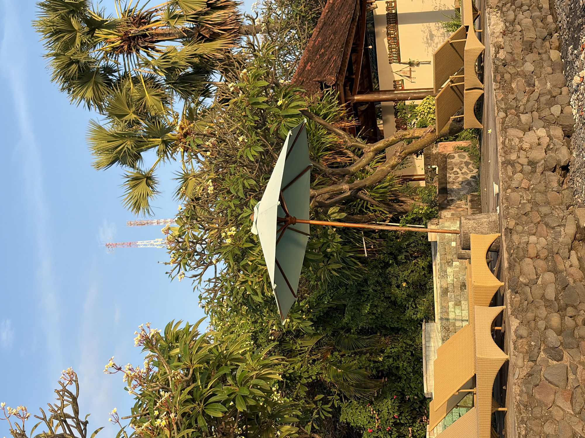 The image captures a serene outdoor scene, likely in Abang, Indonesia, with a focus on a poolside or terrace area. The foreground is dominated by a rough-hewn stone wall, transitioning to a paved area where two tan wicker lounge chairs are positioned. Behind the lounge chairs, a large, light blue umbrella with a wooden pole stands open, providing shade.
The background is lush and tropical, featuring dense green foliage, flowering trees with white blossoms, and several tall palm trees with fronded tops. A rustic, open-air structure with a dark, tiled roof is visible to the right, and a section of a cream-colored wall with decorative elements runs behind it. In the far distance, two prominent red and white telecommunication towers are visible against a clear blue sky. The lighting suggests it is either early morning or late afternoon, with warm sunlight casting long shadows and highlighting the textures of the stone and wicker. No people are present in the scene, contributing to a sense of peaceful solitude.