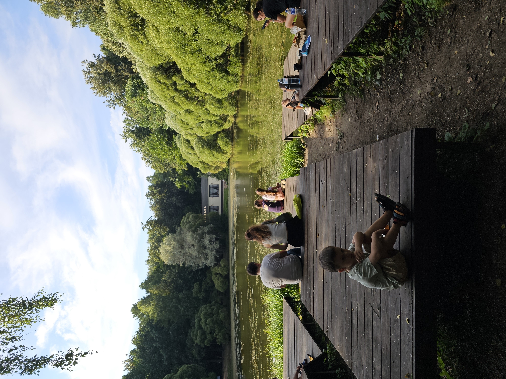 In Moscow, Russia, a wooden pier extends over a tranquil lake within a lush green park. On the pier, a diverse group of people are gathered, some enjoying picnics while others simply relax by the water. In the foreground, a young boy sits on the edge of the pier, his gaze directed away from the camera, appearing contemplative. The scene is bathed in the warm glow of natural sunlight, highlighting the vibrant foliage that encircles the lake and the weathered texture of the wooden planks. The water's surface is speckled with a light green algae or moss, adding to the natural ambiance. In the background, a building with a distinctive sign is visible across the lake, suggesting a park amenity or cafe. The overall atmosphere is one of leisure and peaceful enjoyment of nature.