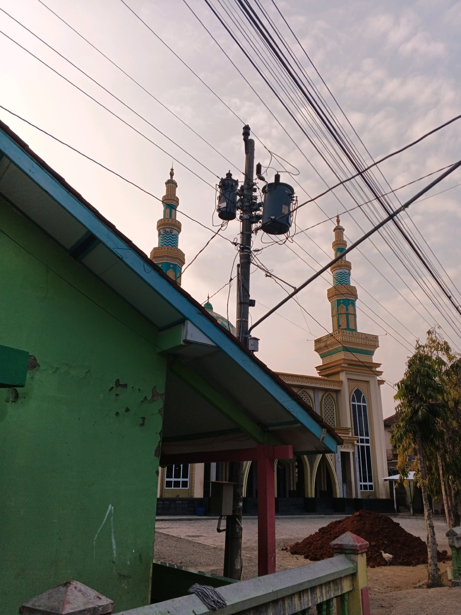 The image depicts a street scene in Kuripan, Indonesia, featuring a mosque with twin minarets in the background. A prominent green building occupies the left foreground, with its peeling paint indicating age or wear. Overhead, a complex network of electrical wires and a transformer on a utility pole crisscross the sky, partially obscuring the view of the mosque. The mosque itself is light-colored with arched windows and decorative details on its minarets, which are topped with spires. In the middle ground, a pile of dark soil sits on the ground, with a small white chicken foraging nearby. Several trees with sparse foliage are visible on the right side of the image. The sky is overcast, suggesting either dawn or dusk. The overall impression is one of a typical Indonesian street scene, with religious architecture, utility infrastructure, and signs of local life.