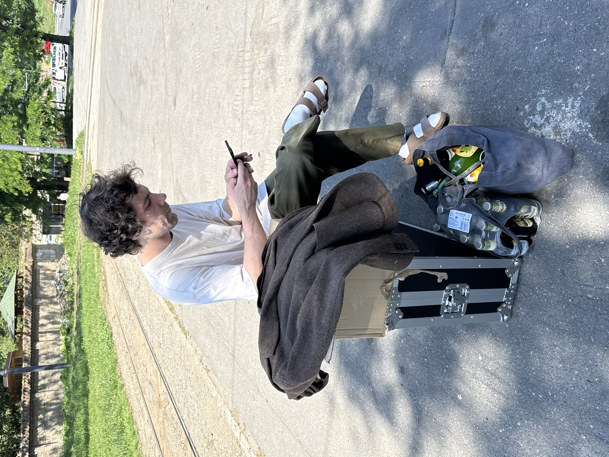 A man sits outdoors on a black and silver hard case atop a cardboard box in Belgrade, Serbia. He has dark curly hair and wears a light-colored t-shirt, green pants, white socks, and brown sandals. He is holding a smartphone in his hands, looking at it. A gray jacket is draped on the hard case beside him. A gray canvas bag and a clear plastic bag filled with bottles, including a bottle of sparkling wine labeled "Schlumberger", sit next to the case. The background shows a park area with trees and grass, and parked cars in the distance. The man sits on a paved surface with tram tracks running alongside.