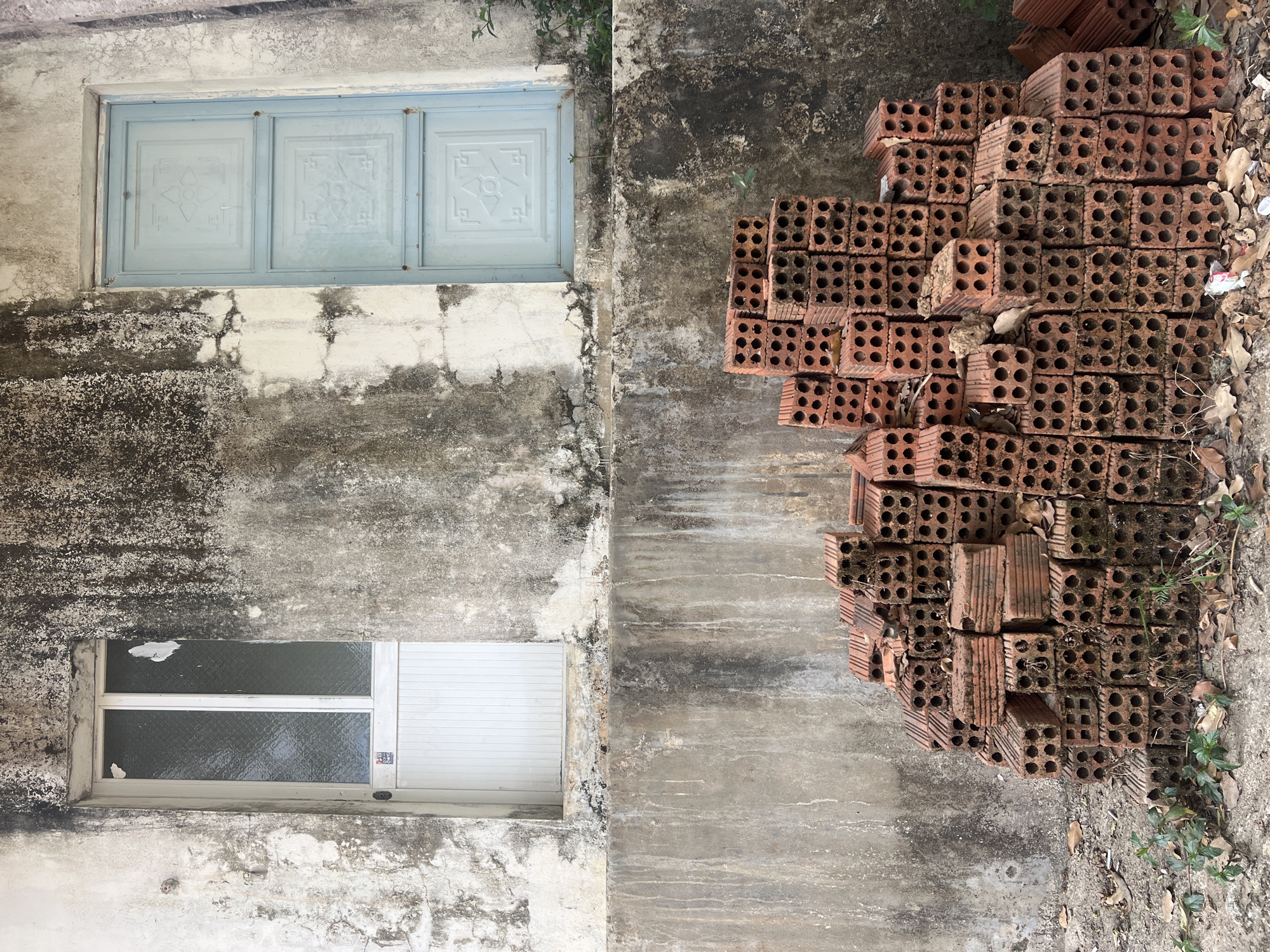 The scene appears to be an exterior shot of a building in Bo Y, Vietnam. The building has a weathered concrete facade with two windows, one with a light blue frame and decorative panels, and another, lower window with a white frame, partially covered by a white sliding door or panel. To the right of the building, there is a large stack of red bricks arranged in an organized yet slightly haphazard manner. The bricks are full of holes. Leaf debris are scattered around the bricks and along the wall of the building.