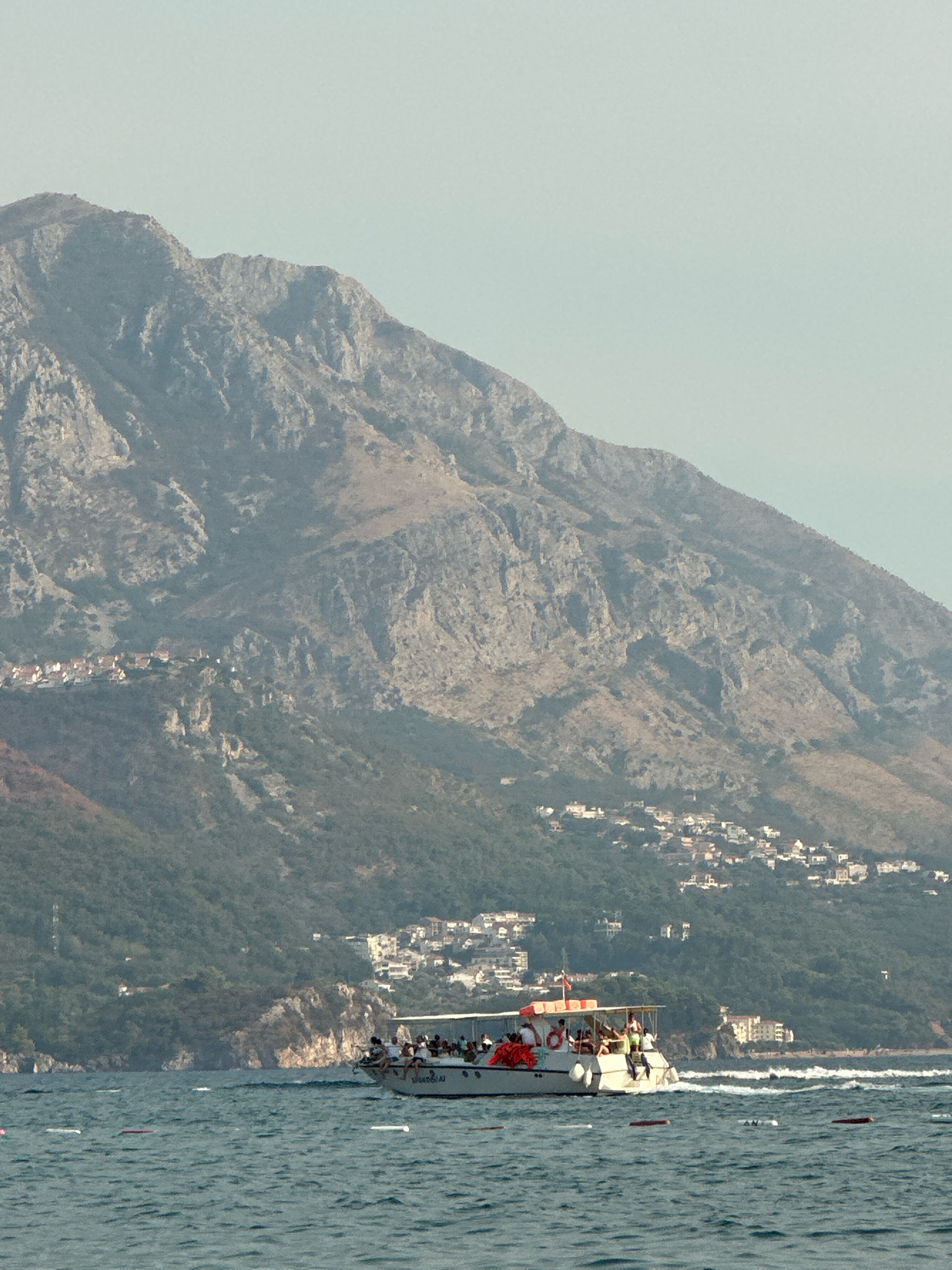 In Rafailovići, Montenegro, a white tour boat carrying approximately 20 passengers sails on choppy blue water. The passengers are dressed in casual summer attire and appear to be enjoying the ride. Some are standing, while others are seated. The boat is positioned in the foreground of the image. In the background, a large, rugged mountain dominates the landscape. The lower slopes of the mountain are covered in dense green vegetation, dotted with white buildings that form a coastal town. The sky is a pale blue, and the overall atmosphere suggests a warm, sunny day.