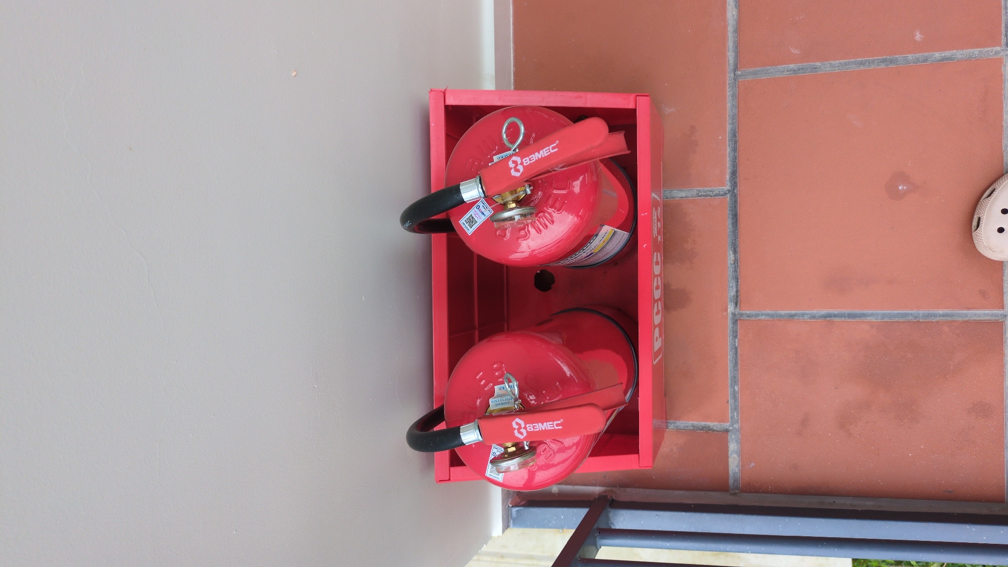 Two red fire extinguishers are stored in a red metal holder. The holder is situated on a floor covered with terracotta tiles. A light-colored wall is visible above the holder. A dark metal railing is present to the left of the holder. The scene appears to be outdoors, possibly on a balcony or patio. The fire extinguishers have black handles and appear to be in good condition. A portion of a light-colored shoe is visible in the bottom right corner of the image. The inscription "PCCC" is visible on the front of the red holder. The overall impression is one of safety equipment being stored in a public or commercial area in Da Lat, Vietnam.