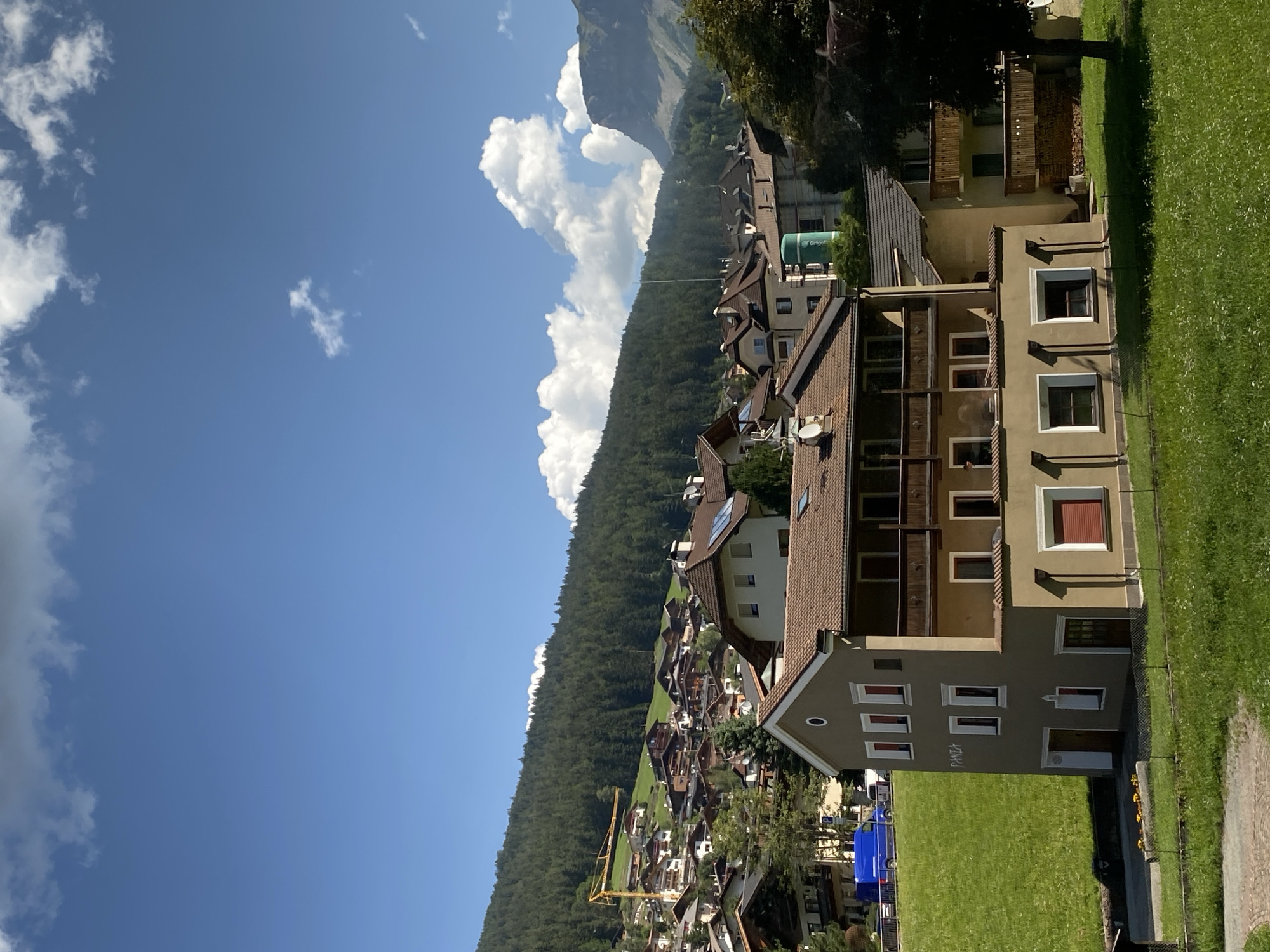 The image shows a view of the town of Urtijëi - St. Ulrich in Gröden - Ortisei, Italy, on a sunny day. In the foreground, a multi-story building with a beige facade and a brown roof is prominent. This building has several windows and balconies, and what appears to be graffiti reading "PIAZZA" on its side. To the left of this building, a lush green hillside slopes upwards, dotted with more houses and a yellow construction crane on the upper slope.
Beyond the foreground building, the town spreads out across the hillside, with numerous houses featuring brown roofs and wooden balconies, typical of Alpine architecture. A dense forest covers the upper part of the hill behind the town. In the background, a majestic mountain peak, likely part of the Dolomites, rises against a bright blue sky with scattered white clouds. The overall scene depicts a picturesque mountain village bathed in sunlight.