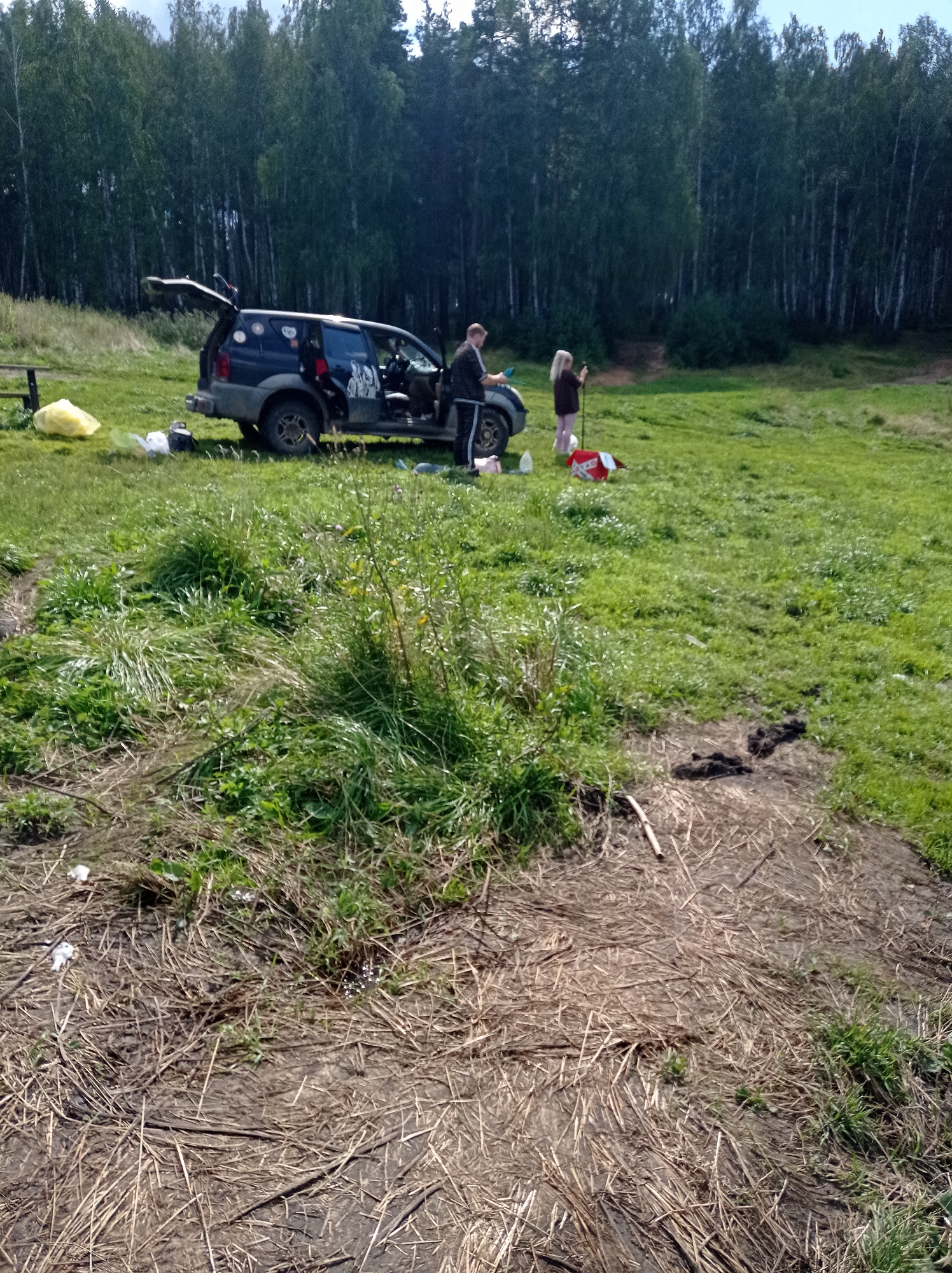 An outdoor scene in the city of Degtyarsk, Russia, shows a dark blue SUV parked on a grassy area with a dense forest in the background. A man in a dark jacket and pants is standing next to the open driver's side door of the SUV, looking towards the rear. A woman with blonde hair, wearing a dark top and pink pants, is standing near the rear of the SUV holding a long pole. A red and white object, possibly a bag or blanket, is on the ground near her. There is a yellow plastic bag to the left of the vehicle, and other miscellaneous items are scattered on the grass. The ground in the foreground is covered in dry grass and some dark soil with hoof prints. The overall scene suggests a stop or activity in a natural setting.