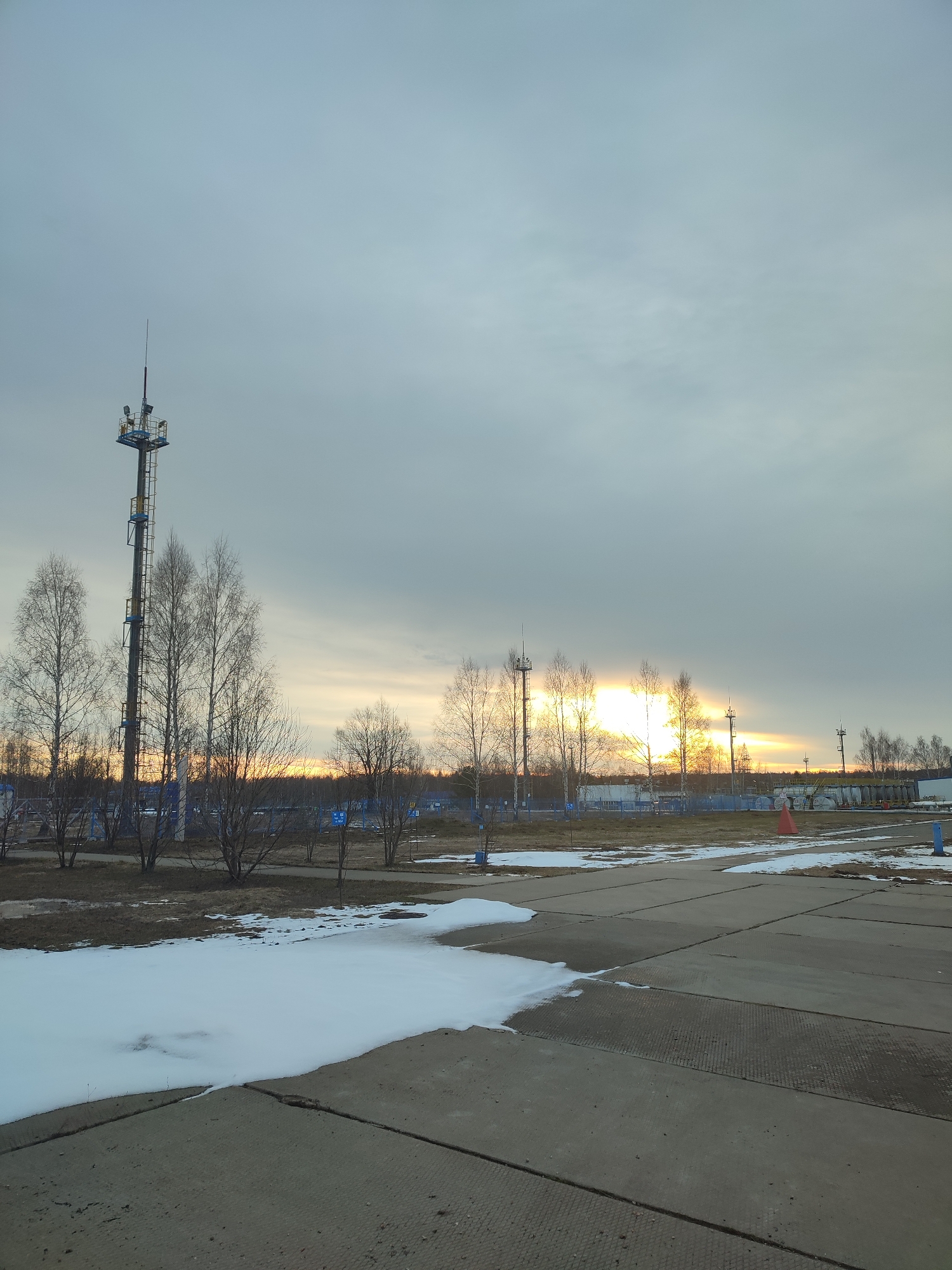 The image shows a partially snow-covered landscape under a cloudy sky, possibly at dawn or dusk. In the foreground, there is a gray concrete surface composed of rectangular sections, with patches of melting snow along the edges. Behind the concrete area, there's a grassy patch with scattered trees, mostly bare of leaves. A tall metal tower with attached equipment stands among the trees to the left. A low blue fence runs across the middle ground, separating the foreground from a more distant industrial area. In the background, a muted sunset or sunrise is visible through the trees. The location is in the city of Unknown, Unknown.