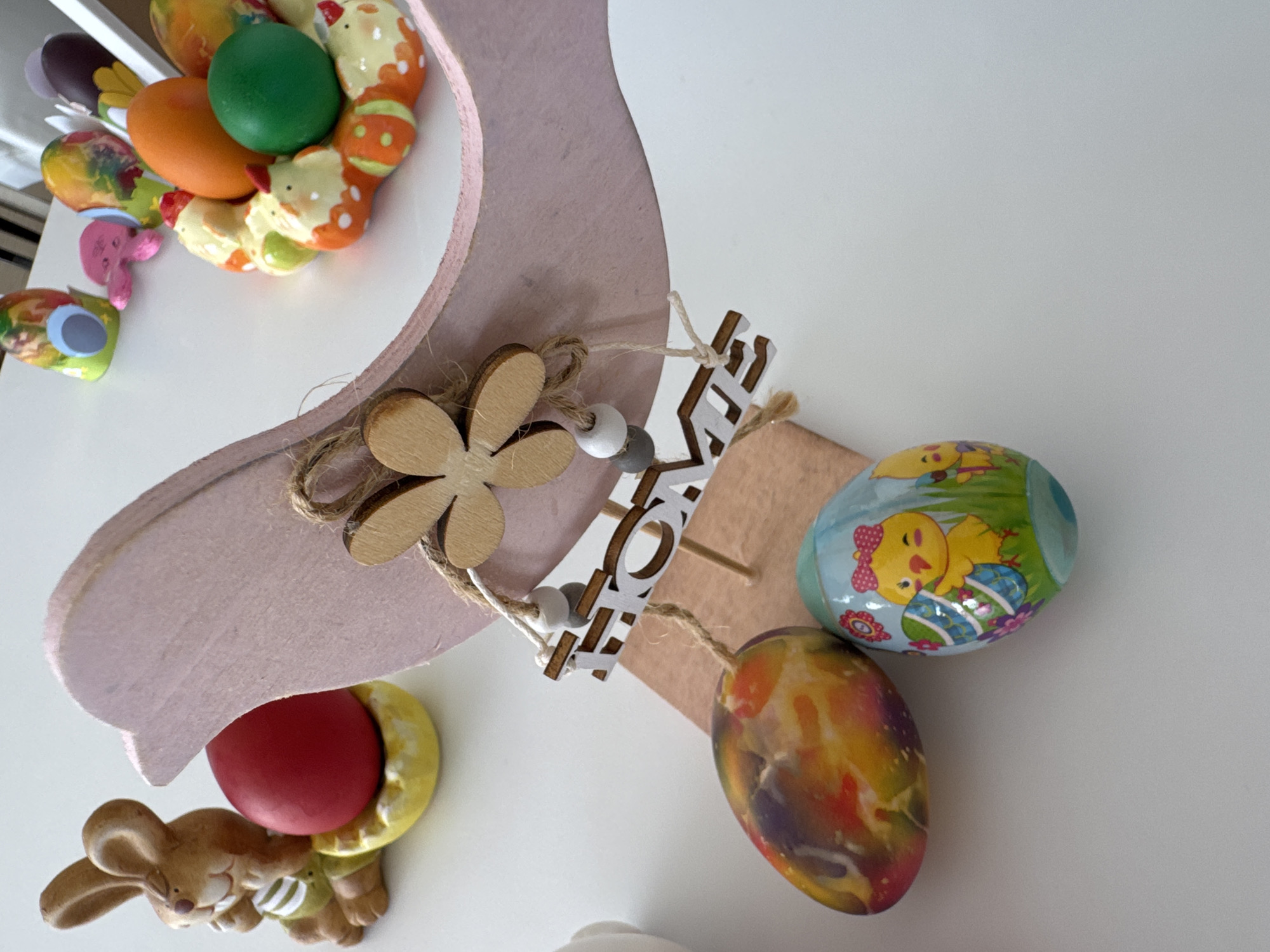 The image shows a close-up of Easter decorations on a white surface. The decorations include a light pink wooden cutout shaped like a rabbit head, decorated with a small wooden flower, a 'HOME' sign made of wood, and two decorated Easter eggs. One egg is blue with a chick design, and the other egg is colored with a marble effect of red, orange, and purple. At the bottom left, there is a rabbit figurine holding a red egg. In the upper left corner, there is a cluster of colorful Easter eggs.