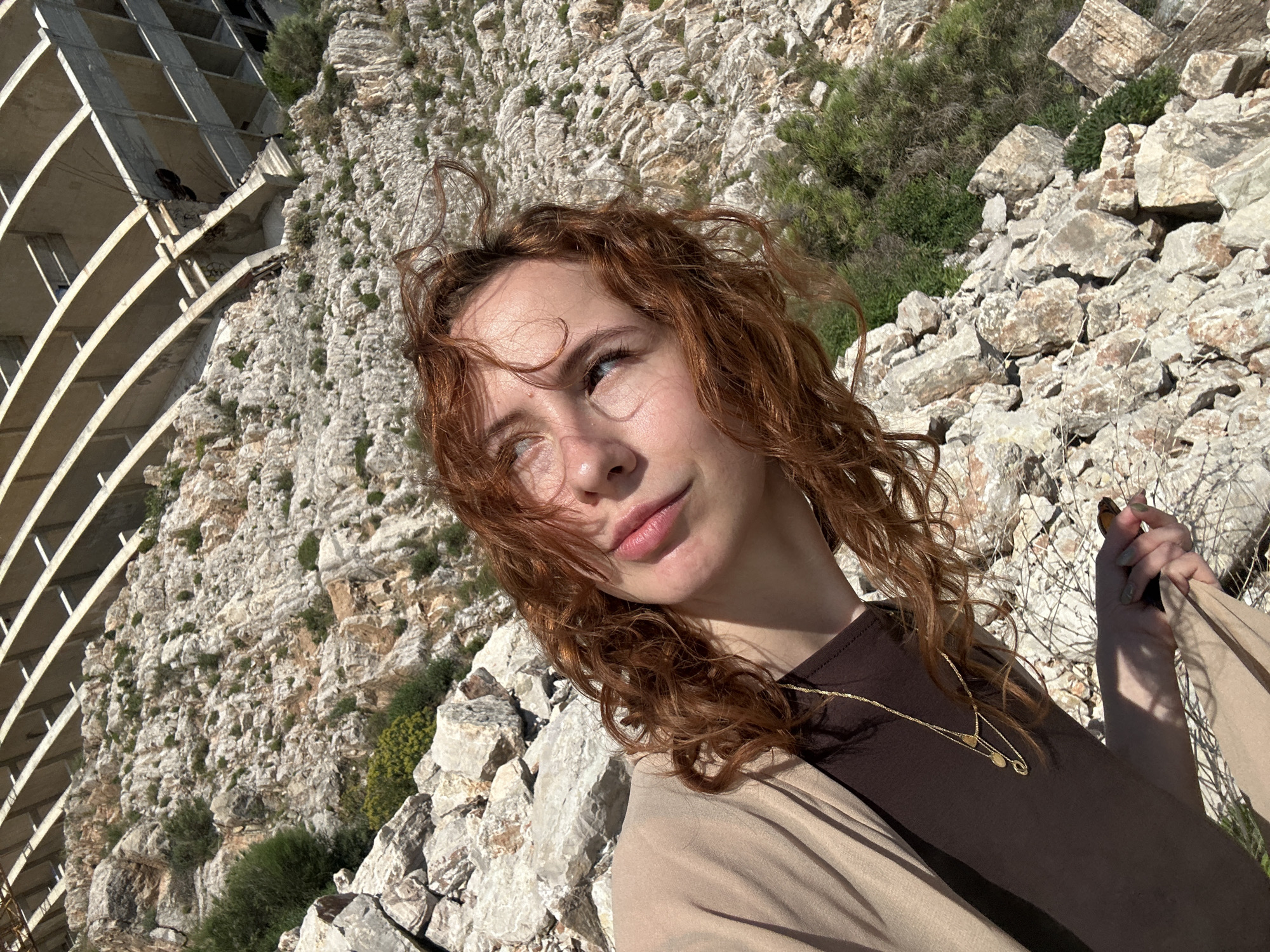 A young woman with red curly hair is captured in a photograph taken outdoors in Perazića Do, Montenegro. She's wearing a brown shirt, a beige jacket, and a gold necklace with two pendants. Behind her, a rugged, rocky landscape slopes upward, dotted with sparse greenery. To the left, the stark, unfinished concrete structure of a multi-story building stands out against the natural backdrop. The photograph appears to have been taken during the day under natural light. The woman is looking slightly upwards and to the left, appearing thoughtful or introspective.