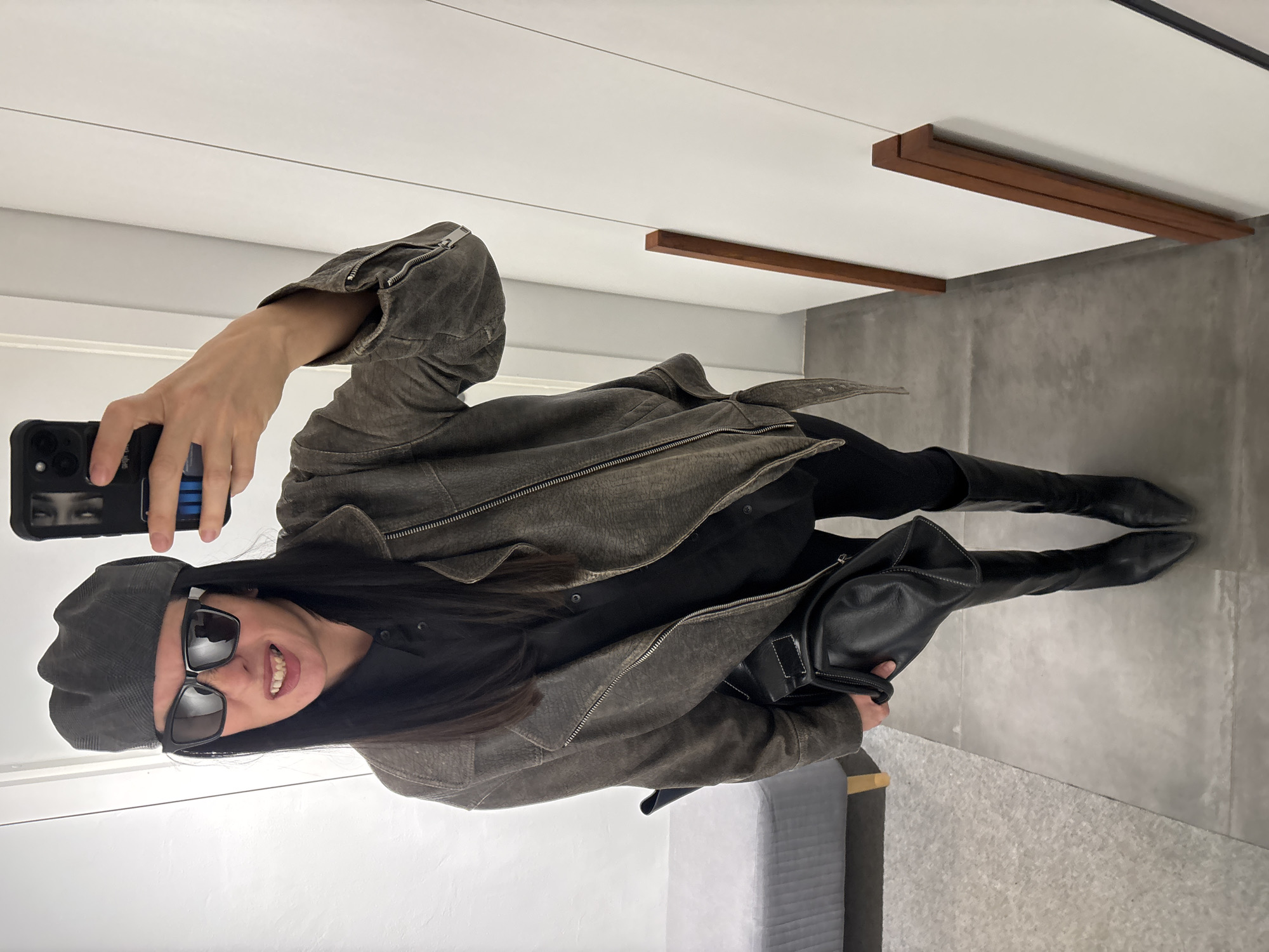 The image is a selfie taken in what appears to be a showroom or upscale retail space. The person taking the photo is a woman wearing a gray beret, large black sunglasses, a textured, gray-brown jacket, black pants, and black knee-high boots. She is holding a phone in her right hand, visible in the upper left corner of the image. The phone case is black with what looks like an image of eyes on it. She is also holding a black leather bag.
The environment features neutral tones with light gray concrete flooring and white walls. There are minimalist wooden fixtures on the ceiling. A gray sofa is visible in the lower right corner.
There is no information available to suggest the image was taken in Chaikovsky, Russia. The location is an interior space with minimal clues as to its specific geographic location.