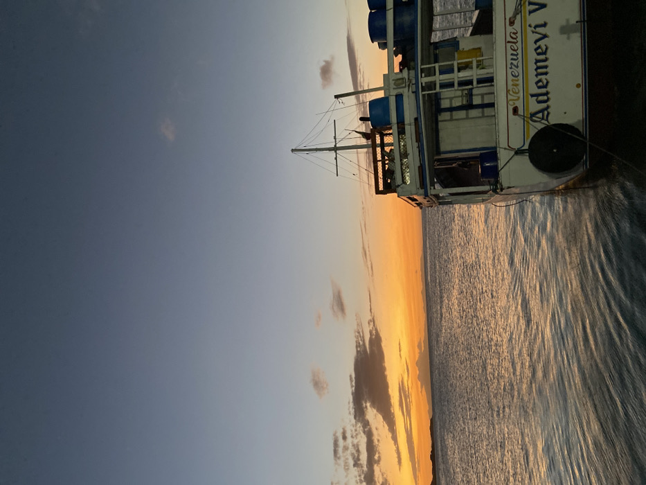 A sideways view of a ship at sea during sunset. The vessel, marked "Venezuela Ademi V", is partially visible on the right. The sea reflects the orange hues of the setting sun, while the sky transitions from a light blue to a dark, cloudless blue. A mast extends upwards from the boat.