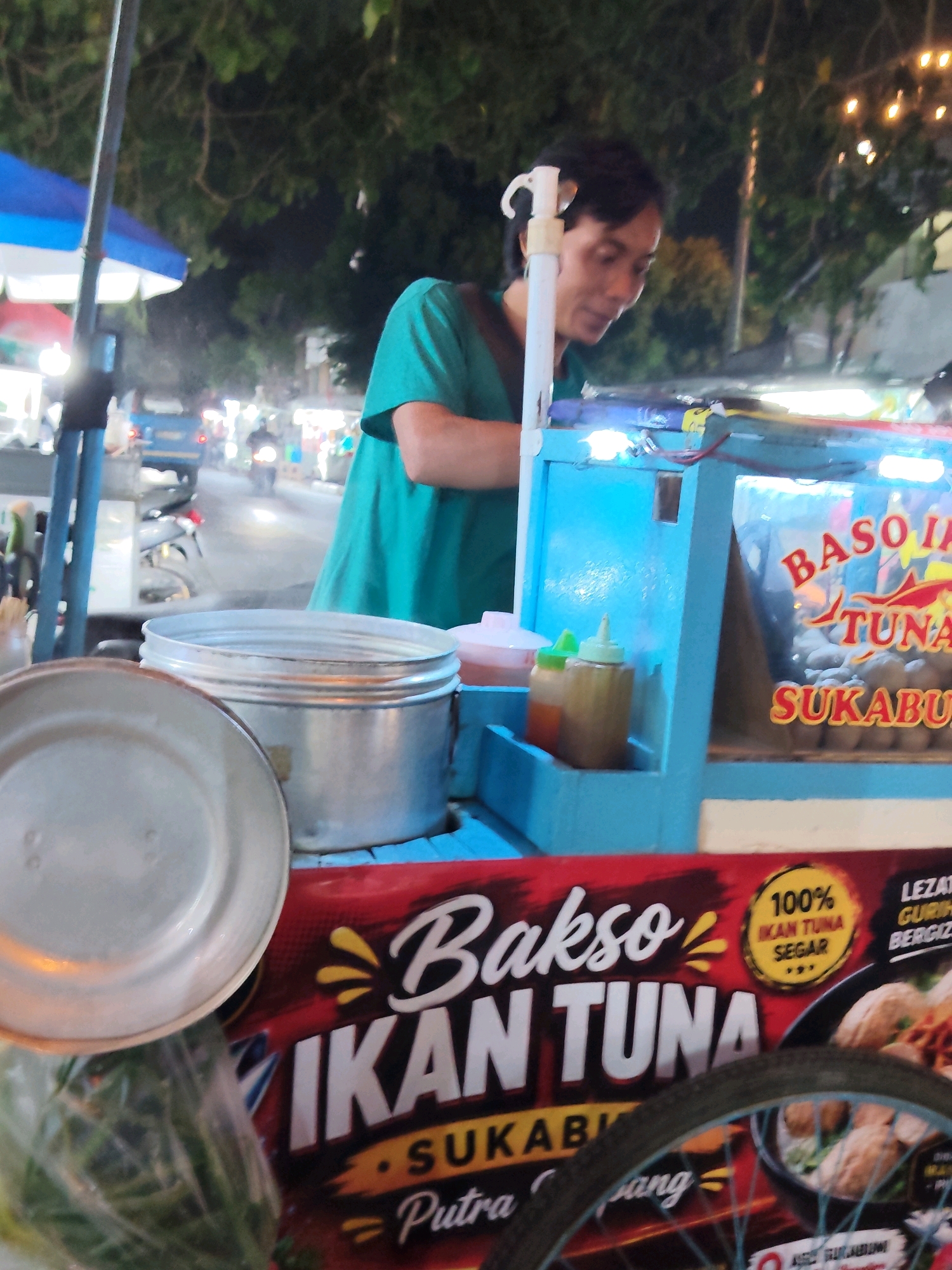 The image shows a street food vendor in Purwakarta, Indonesia. A person wearing a turquoise shirt is preparing food at a cart labeled "BASO IKAN TUNA SUKABUMI". The cart has a blue structure with clear panels displaying meatballs and signage. Two condiment bottles are visible on the cart. In the foreground, a metal plate and a bag of green vegetables are placed on a red surface. In the background, a street scene with vehicles and trees is dimly lit by streetlights, suggesting it is nighttime.