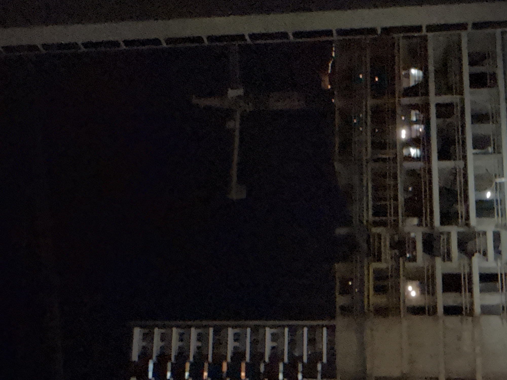 The image shows a nighttime view of a construction site in Tbilisi, Georgia. The scene is dominated by a dark, unlit expanse, likely the night sky or an unlit area of the construction. On the right side of the frame, a partially constructed building stands. It has a grid-like structure with visible beams and concrete elements. Some windows or openings in the building are illuminated, suggesting lights are on inside or from nearby structures. There are reflections in some of these openings, creating bright spots of light.
In the middle ground, a portion of a crane is visible. It appears to be a tower crane, a common sight at construction sites, with its characteristic horizontal arm. The crane is silhouetted against the dark background.
In the foreground, at the bottom of the image, there is a section of what looks like a railing or a fence, with repeating vertical and horizontal elements. Some faint lights are visible through this structure. The overall impression is one of an active construction site at night, with work possibly ongoing or lights left on. The image is grainy, likely due to low light conditions and camera settings. No people are clearly discernible in the image. The time of day is clearly night.