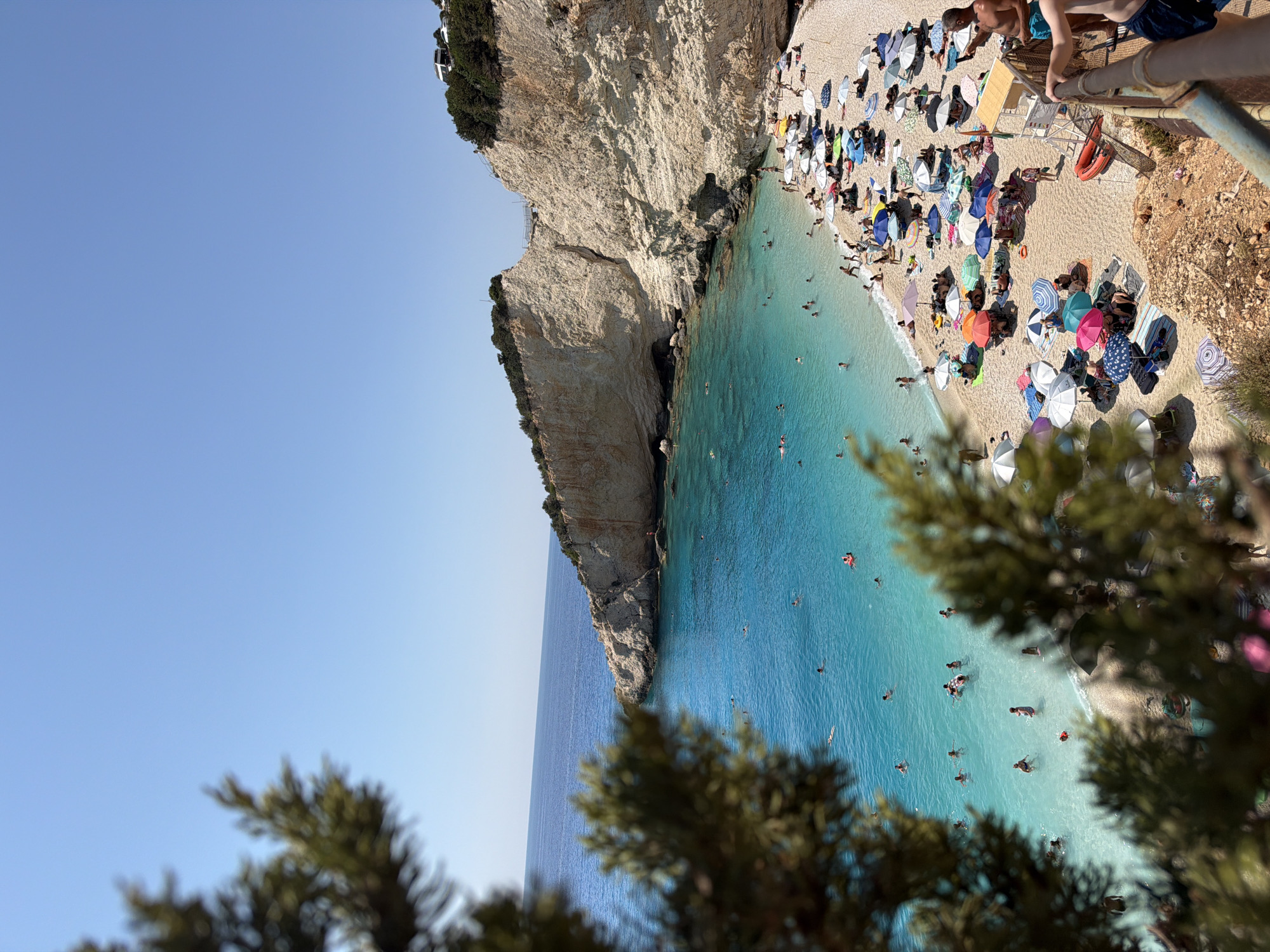 This is a high-angle, full shot of a crowded beach on a sunny day. The beach is located in the Municipal Unit of Apollonioi, Greece, likely Egremni Beach on the island of Lefkada, known for its white cliffs and turquoise waters. The foreground is partially obscured by the dark green foliage of trees. The main scene unfolds on a wide, white pebble beach teeming with people relaxing under numerous colorful beach umbrellas and parasols. Many individuals are swimming and wading in the crystal-clear, bright turquoise water, some on inflatable rafts. The backdrop is dominated by towering, sheer, light-colored cliffs that frame the bay, with a dense green canopy of trees and some buildings visible at the top of the cliff on the right. The sky is a clear, deep blue. A metal railing is visible in the lower right corner, suggesting a viewpoint or walkway. The overall impression is one of a popular tourist destination experiencing a busy summer day.