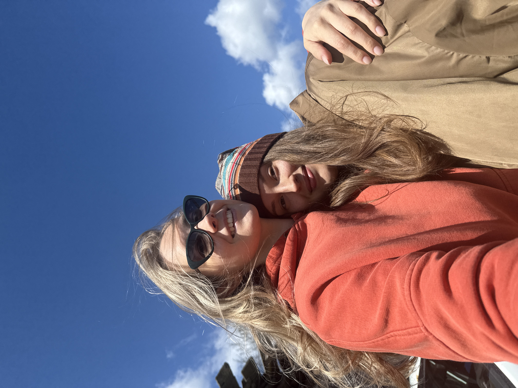 Two people are posing for a selfie under a clear blue sky with a few white clouds. The person in the foreground has blonde hair and is wearing sunglasses and an orange hoodie. The person in the background has long brown hair and a colorful beanie. They are close together, possibly embracing. The location is Chaikovsky, Russia.