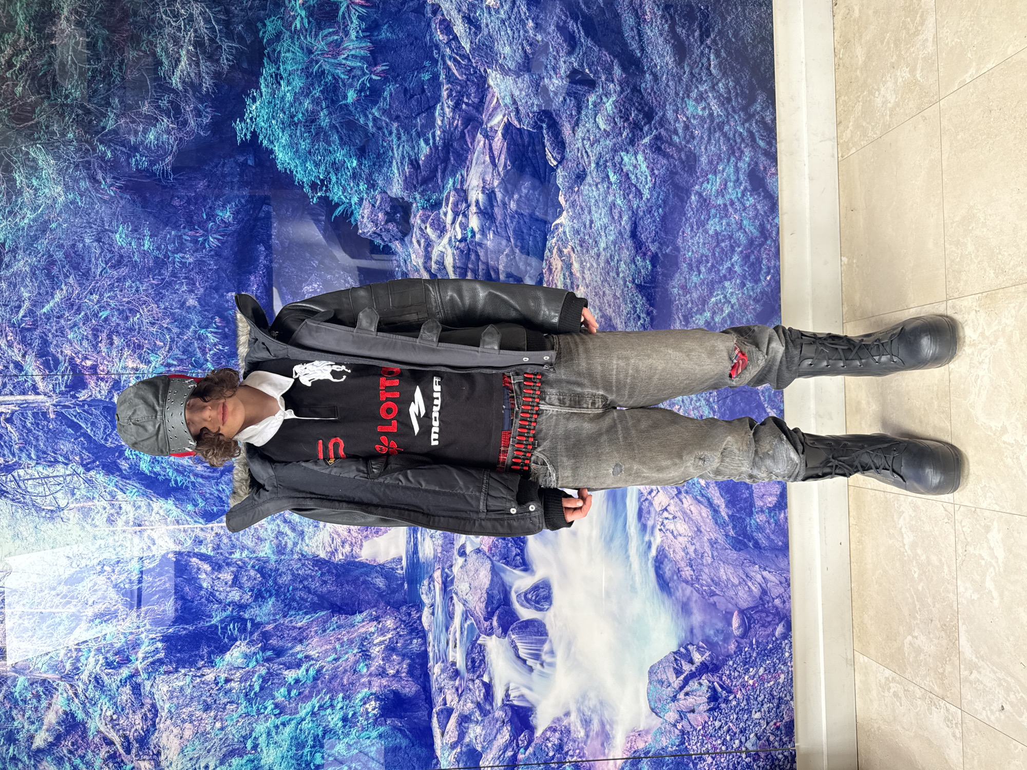 The image shows a person wearing a cap, black jacket, black shirt with the word "LOTTO" printed on it in red and white, gray jeans with some tears and red patches visible, and black boots. They are standing in front of a wall with a blue and white mural that resembles a landscape with rocks and water. The floor is tiled with light-colored tiles. The location is Unknown, Unknown.