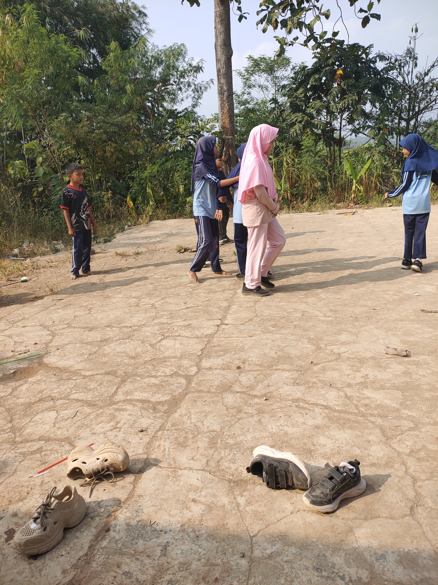 In Subang, Indonesia, an outdoor scene unfolds on a sun-drenched, cracked concrete ground. Several children are present. On the left, a young boy in a black t-shirt with a red collar and dark pants stands looking to the left. Further in the center, three children, two wearing headscarves (one pink, one blue), and another in a blue hijab and light blue shirt, appear to be engaged in an activity, possibly a game, with one child facing away from the camera. To the far right, a child in a blue hijab and light blue shirt stands facing the group in the center, with their right hand extended. In the foreground, resting on the concrete, are several pairs of shoes: two light-colored, open-toed sandals, and a pair of dark, velcro-strap sneakers. The background is a dense expanse of lush green trees and vegetation.